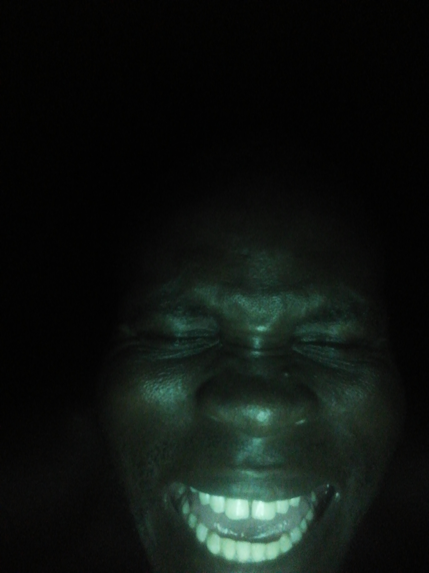 The image depicts a close-up of a person's face in dark lighting. The person has dark skin, closed eyes, and a wide, toothy smile. The overall tone of the image is green.