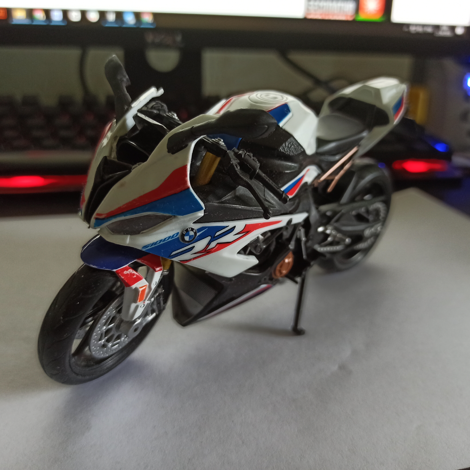 A close-up, slightly angled shot shows a detailed model of a white, red, and blue BMW S1000RR motorcycle. The motorcycle is positioned on a white surface, with a blurred background of computer monitors and a keyboard that is lit with red and blue lights. The model is highly detailed, featuring realistic tires, suspension, and bodywork, including the BMW logo and S1000 branding. The lighting suggests it is indoors, possibly in a home or office setting. There is no information in the image to indicate the location in the city of Nizhny Novgorod, Russia.