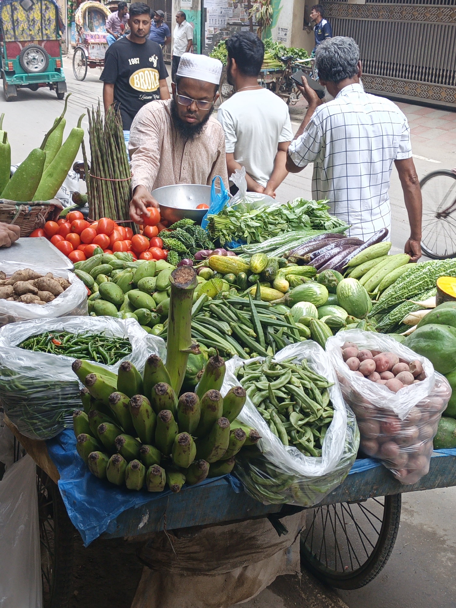 This image captures a bustling street market scene in Dhaka, Bangladesh, focusing on a mobile vegetable and fruit stall. The central figure is a male vendor wearing a white prayer cap, glasses, and a light-colored patterned shirt, seen reaching for a red tomato with his right hand above a metal bowl. He has a dark beard.
The market stall, a blue-painted wooden cart with bicycle wheels, is overflowing with a wide variety of fresh produce. In the immediate foreground and center, a large bunch of green, unripe bananas is prominently displayed. Surrounding these are numerous other items: piles of bright red tomatoes, bags of green chilies, bags of small red potatoes, and large quantities of ginger or smaller potato-like roots. Various green vegetables are meticulously arranged, including elongated gourds (like bottle gourds or calabash), ridge gourds, pointed gourds, bitter gourds, okra, green mangoes, and different types of eggplants (both round green and long purple varieties). Bundles of green leafy vegetables and long green beans (possibly drumsticks or yardlong beans) also cover the cart.
Several other people are visible in the background and surrounding the stall. Behind the vendor, a man in a black t-shirt with the text "1987 BROKE" (and partially visible "Leisure Club" below) stands observing. To the right, another man, viewed from behind, wears a white-and-black plaid shirt and holds a black smartphone to his ear. In the further background, a green auto-rickshaw and a traditional cycle rickshaw are seen on a paved street lined with buildings, characteristic of a South Asian urban setting. The scene appears to be taking place during the day, under natural light, depicting a lively daily market activity.