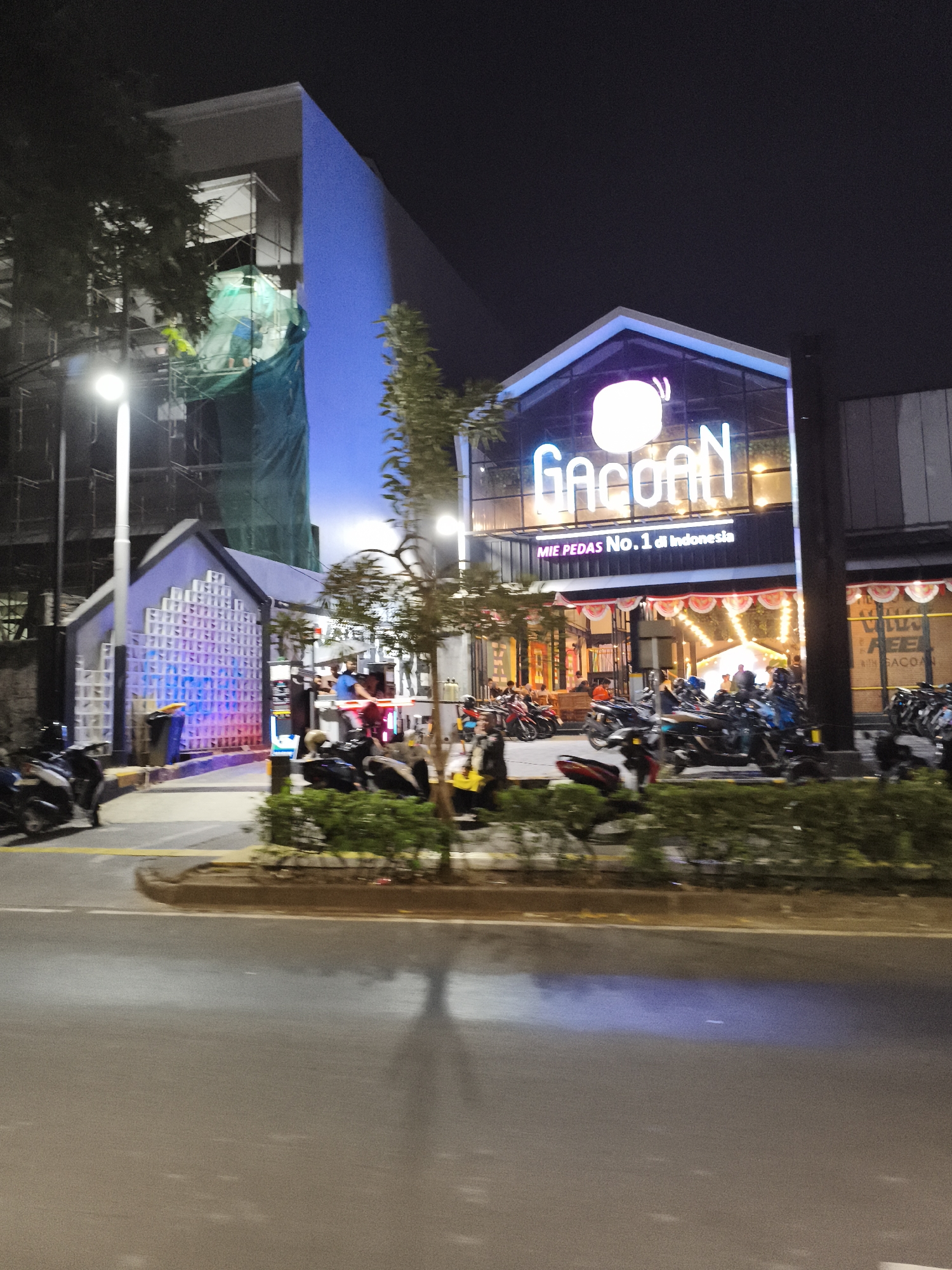 The image depicts a street scene at night in South Jakarta, Indonesia, featuring a brightly lit establishment with a neon sign that reads "Gacoan." The establishment appears to be a restaurant, with "MIE PEDAS No. 1 di Indonesia" (Spicy Noodles No. 1 in Indonesia) written below the main sign. In front of the establishment, there is a parking area with numerous motorcycles. The building itself has a modern architectural style with large glass windows. Adjacent to the restaurant, a building is under construction, covered with green scaffolding and netting. The street in the foreground is wet, reflecting the lights from the surroundings. Several people are visible, some standing near the entrance of the restaurant and others appearing to be working on the construction site. The overall atmosphere is urban and lively, characteristic of a commercial area at night.