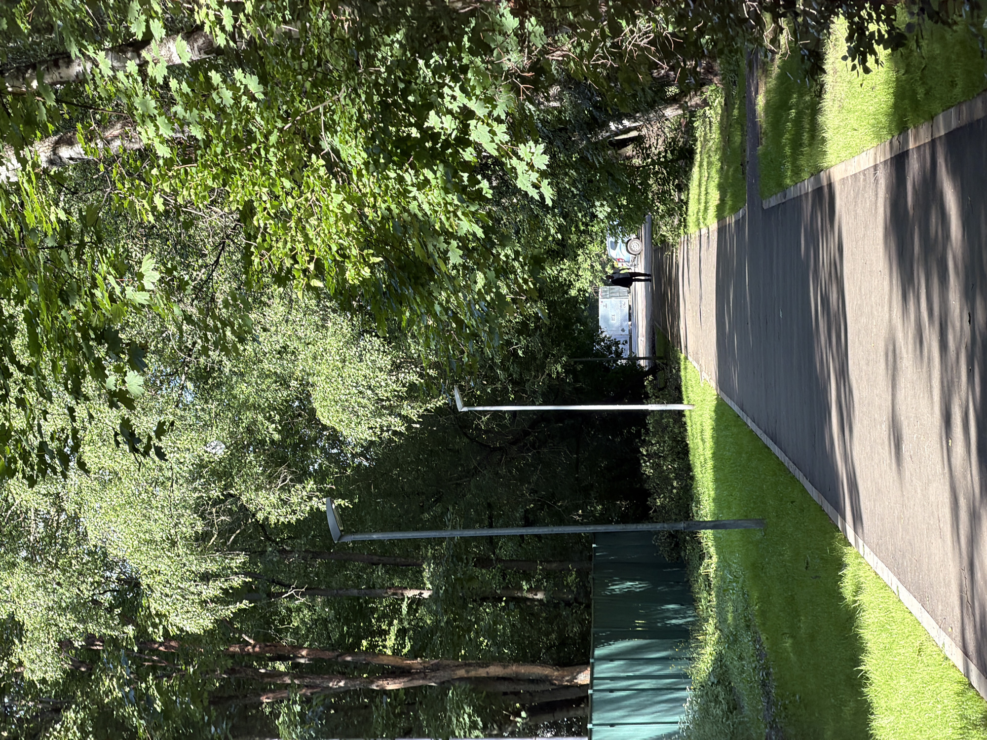 This is a full-shot, eye-level, outdoor, vertical photograph of a pathway bordered by lush green grass and trees. The pathway appears to be paved with asphalt and has a light-colored border. Sunlight filters through the dense green foliage of the trees, casting dappled shadows across the path. On the left side of the path, a tall, dark green fence is visible. Two modern, minimalist streetlights stand along the left side of the pathway. In the distance, a person is walking away from the camera towards a light-colored vehicle and a large, white, modular structure. The scene is set in the city of Khimki, Russia, likely in a park or residential area given the presence of the pathway, manicured grass, streetlights, and fencing.