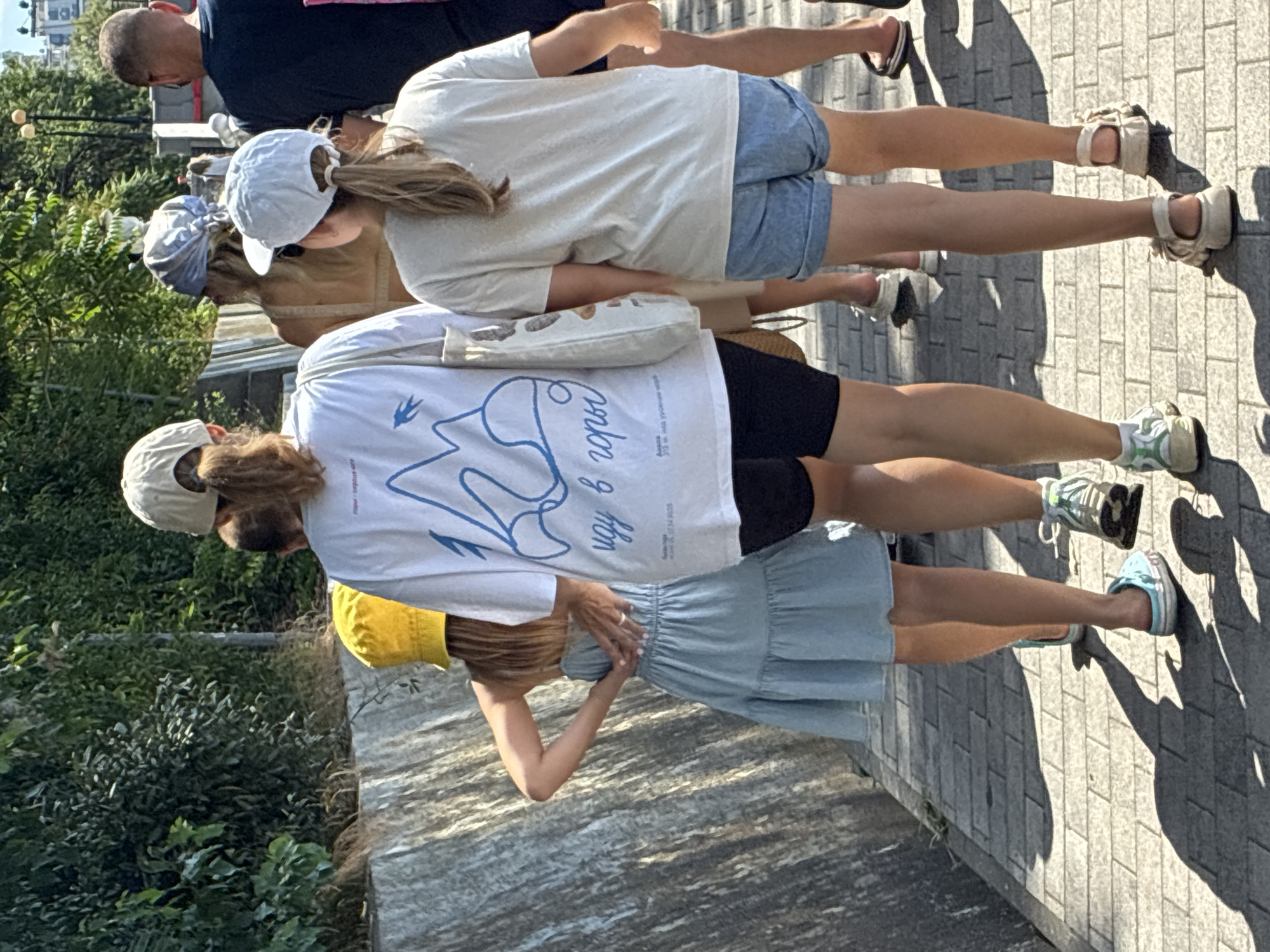 The image shows a group of people walking on a brick pathway in Sochi, Russia, on a sunny day. A woman in a white t-shirt with a blue mountain graphic and text, black shorts, and a cap is in the foreground, facing away from the camera. To her left, a young girl in a light blue dress and a yellow bucket hat walks with her hands behind her back. To the right of the woman, a young girl in a white t-shirt and denim shorts walks with her hands at her sides. Behind them, other individuals are visible, including a person in a beige dress and a man in a dark t-shirt. The pathway is lined with lush green foliage on the left and a stone retaining wall. The shadows cast on the pathway suggest it is midday.