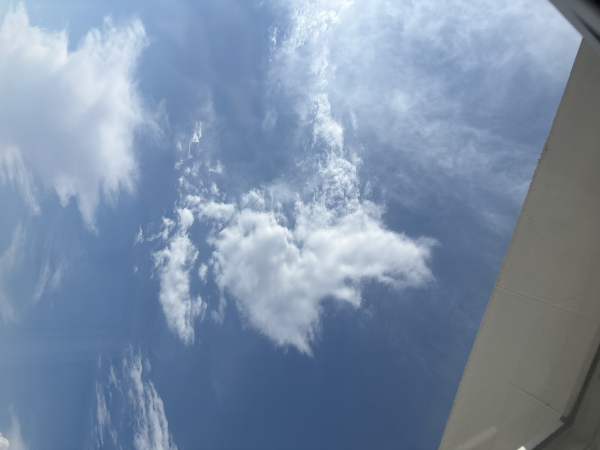 The image depicts a view of the sky from an elevated position, possibly from an airplane or a tall building. The sky is predominantly blue with scattered white clouds of various shapes and sizes. Some clouds are wispy and thin, while others are larger and more voluminous, with some appearing fluffy and textured. In the upper portion of the image, a portion of a white, angular structure is visible, likely a wing of an aircraft or part of a building's exterior, suggesting an urban or aerial perspective. The lighting indicates it is daytime, with the sun likely casting light from the left, creating subtle shadows and highlights on the clouds. There are no people or discernible landmarks of Засечное, Russia visible in the frame. The scene is serene and focuses solely on the atmospheric conditions.