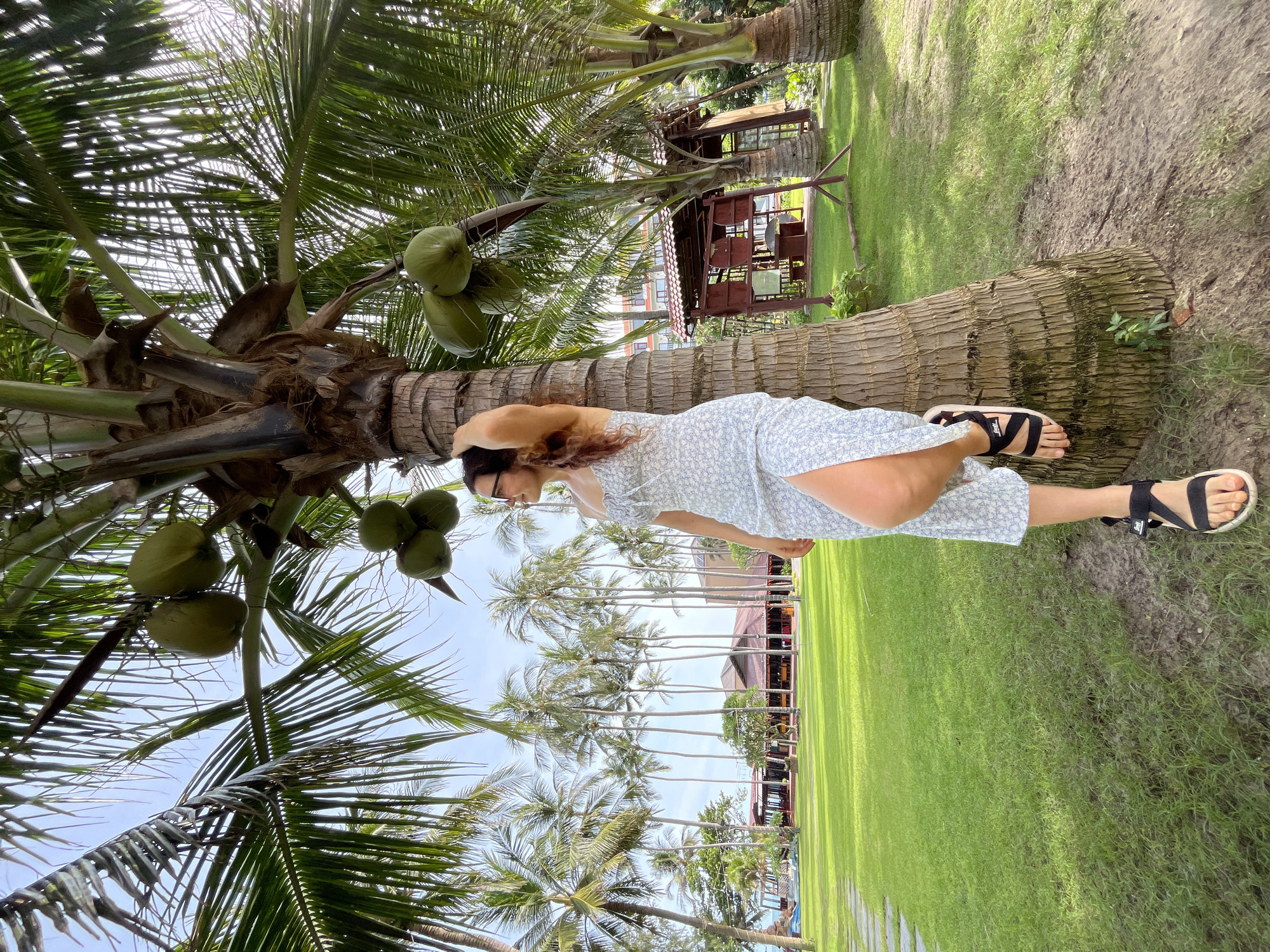 In Mũi Né, Vietnam, a woman with dark, wavy hair, wearing glasses and a light blue floral sundress with a thigh-high slit, stands leaning against a thick palm tree trunk. She has one leg bent and resting against the tree, her hand raised to touch her hair. Her black sandals with white soles are visible. The palm tree above her has several green coconuts. The background features a lush green lawn with a pathway, numerous tall palm trees, and a building with a reddish-brown tiled roof. The sky is bright blue with some clouds. The scene appears to be a resort or a park.