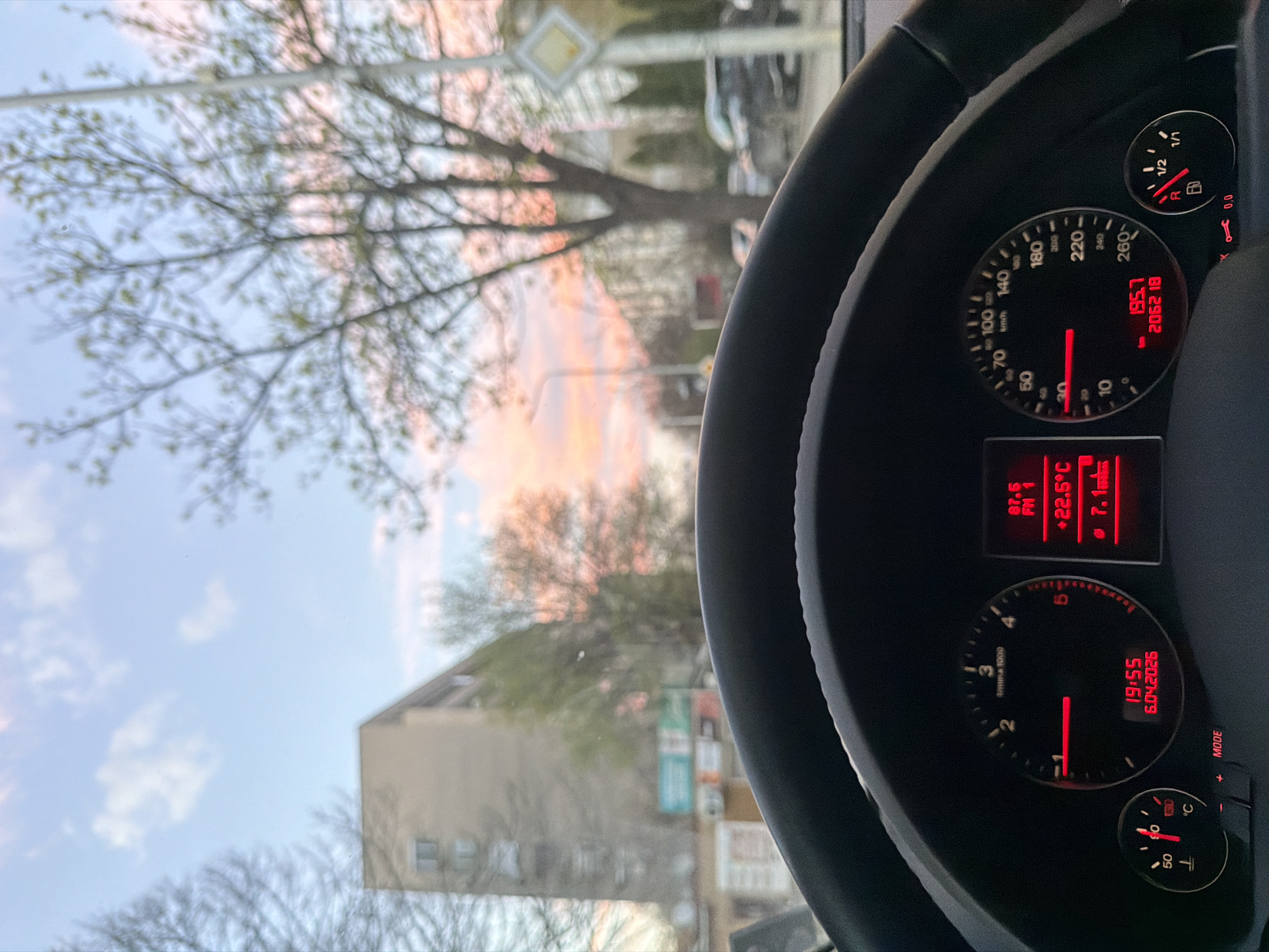 The image is taken from the driver's seat of a car, showing part of the dashboard, steering wheel, and the view through the windshield. The speedometer reads 195.7, and the odometer reads 206218. The dashboard also displays the time as 19:55 on April 6, 2026, an outside temperature of 22.5 degrees Celsius, and the radio tuned to 87.5 FM1. Outside, a street scene is visible with trees, buildings, and a sky with a pink-orange sunset glow. Based on the street sign shape that is vaguely visible behind the dashboard, the location is likely Plovdiv, Bulgaria.