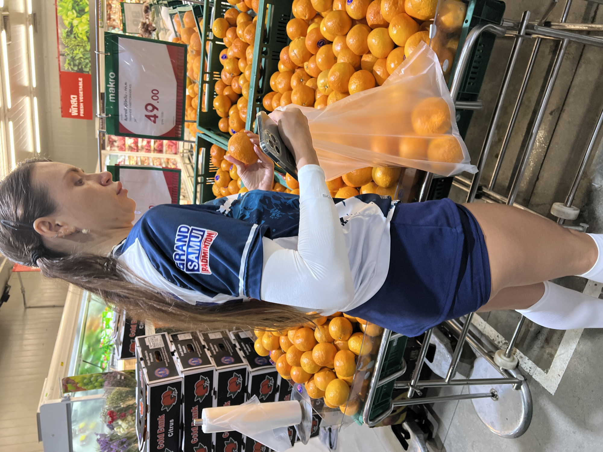 In a brightly lit grocery store aisle, a woman with long brown hair, wearing a navy blue athletic top with "GRAND SAMUI BADMINTON" emblazoned on it and white knee-high socks, is captured from the waist up. She is holding a clear plastic bag filled with oranges and a smartphone, appearing to be in the process of selecting produce. The background shows shelves stocked with produce, including a large display of oranges in green crates and boxes of citrus fruit. A sign above the oranges reads "makro" and "FRESH VEGETABLE" in English and Thai, with a price of "49.00" displayed prominently. The setting appears to be a supermarket in Baan Bo Phut, Thailand, as indicated by the text on the sign and the overall context. The lighting suggests it is daytime, and the indoor setting implies controlled weather conditions. The woman's expression is focused and neutral as she interacts with the fruit and her phone, suggesting a routine shopping activity.