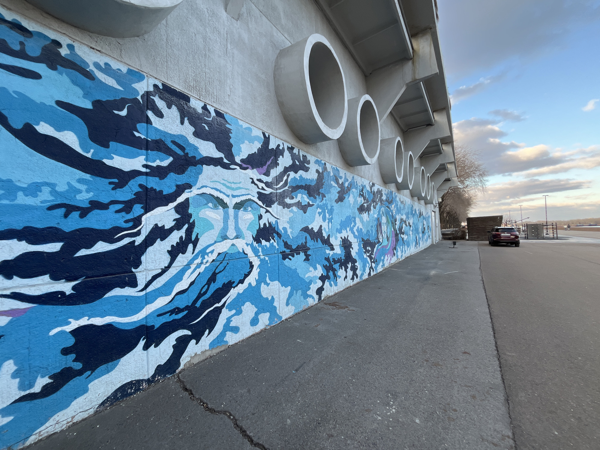 A wide-angle, low-perspective outdoor shot captures a long concrete wall adorned with a vibrant blue and white mural in Krasnoyarsk, Russia. The mural features a stylized, ethereal face, possibly a water spirit or mythical figure, integrated into flowing, wave-like patterns in various shades of blue, white, and dark blue, creating a dynamic, almost camouflage-like effect.
Above the mural, along the upper part of the light grey concrete wall, several large, hollow, circular concrete structures protrude outwards, repeating in a rhythmic sequence towards the horizon. These elements add an industrial or architectural dimension to the scene.
An asphalt path or road stretches alongside the wall in the foreground and midground, showing some cracks. In the distance, a dark red SUV is parked on the road, and tall streetlights are visible. Beyond the wall and its structures, bare trees or bushes can be seen under a partly cloudy sky, with patches of blue visible. The lighting suggests late afternoon or early morning, casting a soft and even illumination across the scene. The overall ambiance is calm and visually striking due to the contrast between the organic mural and the rigid concrete structures. No discernible text is visible in the image.
