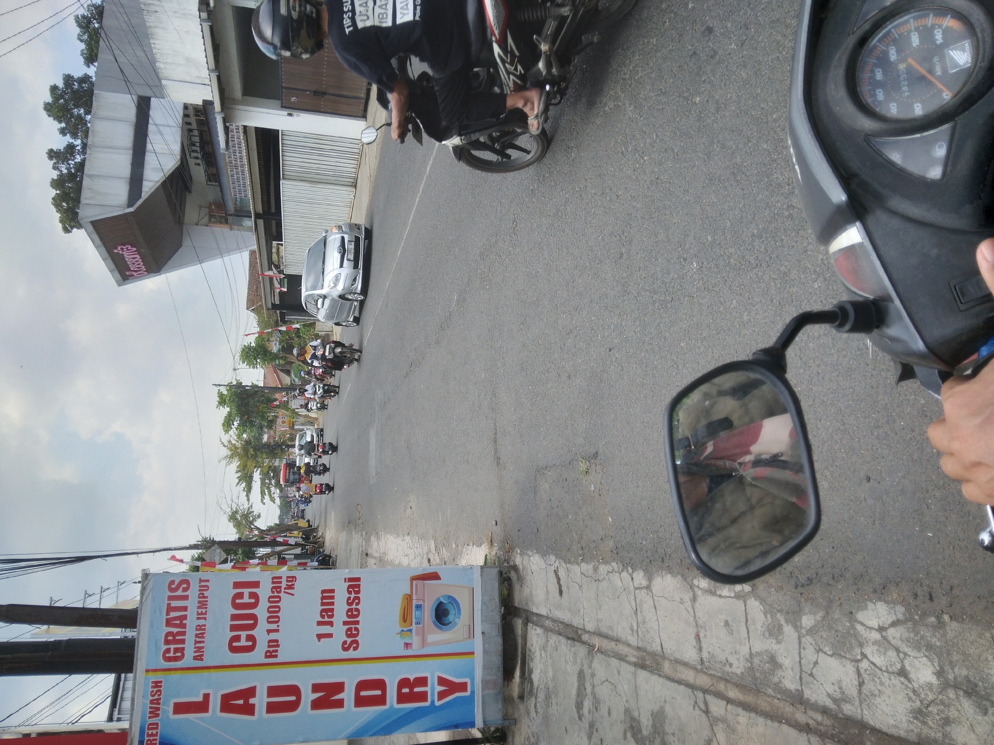 The image depicts a street scene in Purwokerto Wetan, Indonesia, captured from the perspective of someone riding a motorcycle. In the foreground, the rider's hand is visible on the handlebars, and a portion of the motorcycle's dashboard with a speedometer reading is also in view. A side mirror reflects the rider and their surroundings, showing a patterned fabric item hanging.
To the left of the road, a tall blue and white banner advertises laundry services, with the words "RED WASH," "GRATIS ANTAR JEMPUT," "LAUNDRY," "CUCI Rp 1.000an /kg," and "1 Jam Selesai" (1 Hour Complete) prominently displayed. A graphic of a washing machine and folded laundry is also on the banner. The banner is attached to a pole, which is also supporting electrical wires.
The road itself is paved and shows some wear. Several motorcycles are in motion on the road, some carrying one or two riders. A silver car is also present, driving in the same direction as the motorcycle the photo is taken from. In the background, buildings line the street, including a modern structure with a distinctive angled roof and the word "Kassanta" visible on its facade. Trees and utility poles are also part of the urban landscape. The sky is partly cloudy. The overall impression is that of a typical day in a bustling Indonesian town.