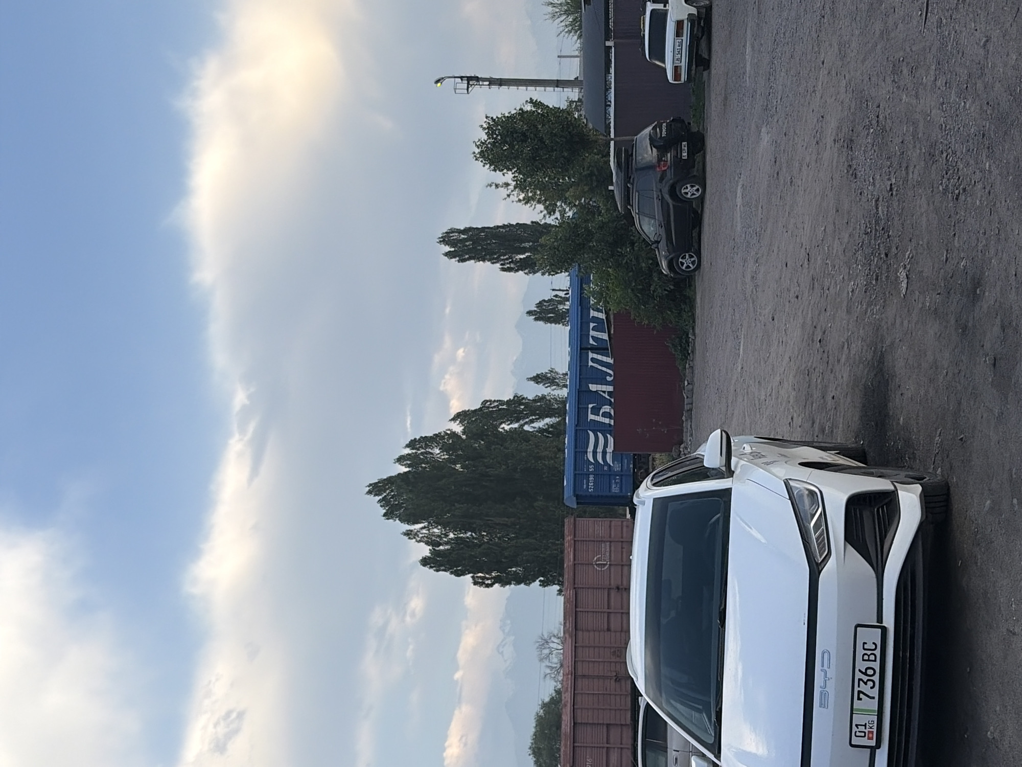 The image shows an outdoor scene with several vehicles and shipping containers. In the foreground, a white car with the license plate "01 736 BC" is parked. Behind it, a dark-colored SUV with "TOYOTA" visible on the rear is parked, followed by a blue shipping container with Cyrillic text that appears to read "БАЛТІ" and some numbers. Further back, a white car is parked. The scene is set in a lot with a gravelly surface, and trees and mountains are visible in the background. The sky is partly cloudy with bright sunlight, suggesting it is daytime, likely late afternoon due to the warm light. There are no people visible in the image. The overall impression is one of an industrial or transportation hub.