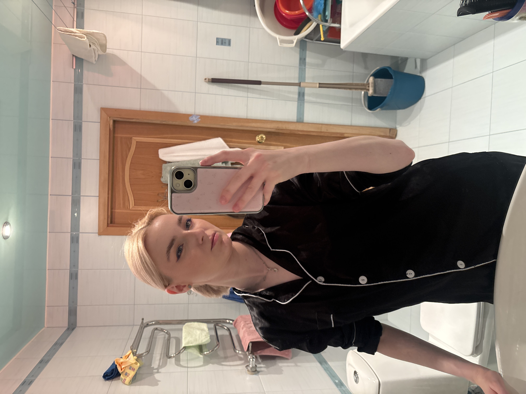 A young woman with blonde hair pulled back in a ponytail, wearing black pajamas with white trim, stands in a bathroom and takes a mirror selfie with her pink phone. She is wearing a necklace and her makeup includes winged eyeliner and blue eyeshadow. The bathroom features white tiled walls, a wooden door, a toilet, a towel rack with towels, a mop and bucket, and a bathtub. The scene is set in the city of Sinyavino, Russia.