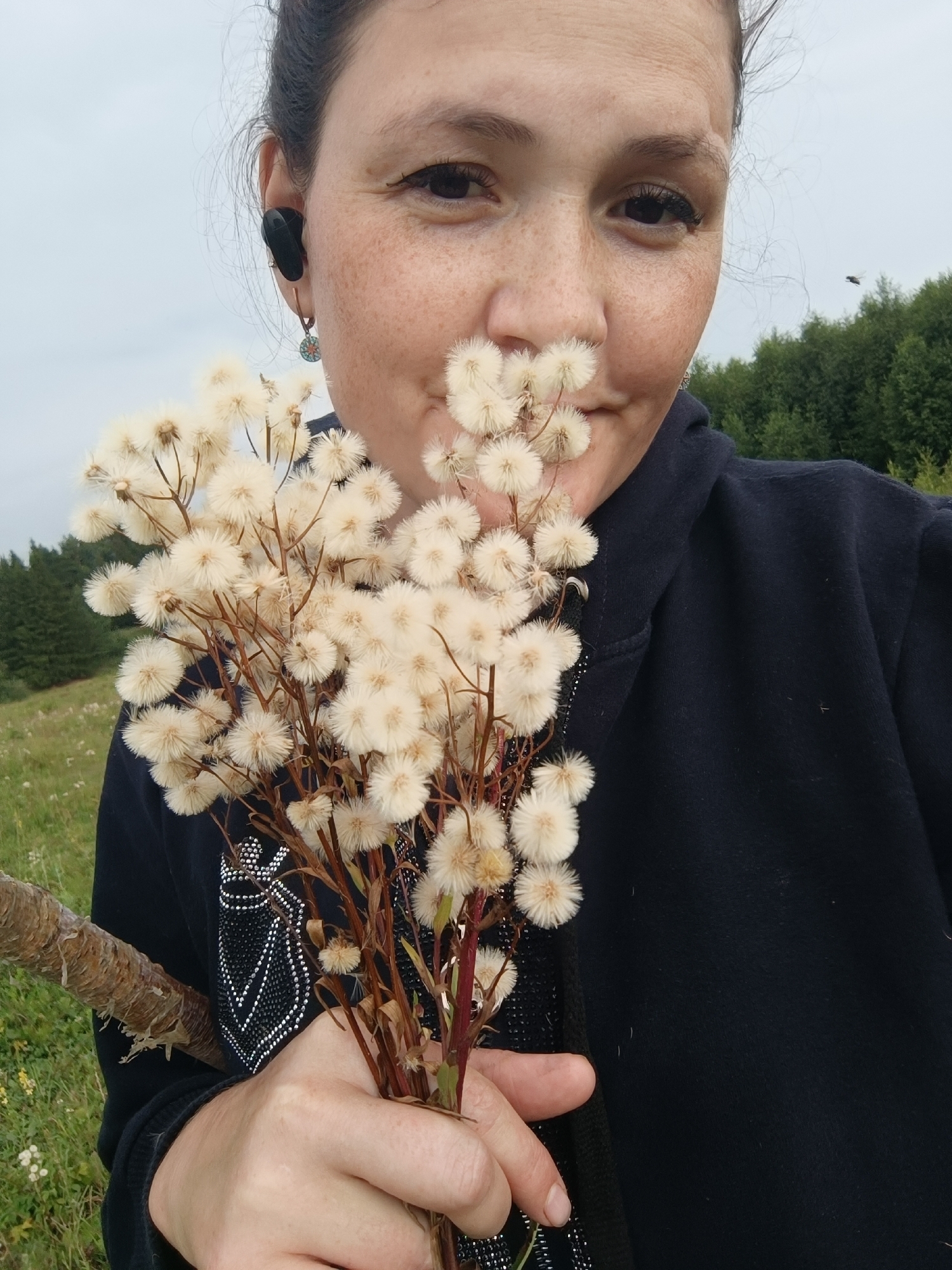 An outdoor, medium shot shows a woman in a black hoodie holding a bouquet of white, fluffy wildflowers near her face. She has light brown skin, dark brown hair pulled back, and is wearing black earbuds and a small, circular earring. She is smiling, and her face is partially obscured by the flowers. The background is a blurry green forest under a cloudy sky. The photograph appears to have been taken in a rural or natural setting. The location in the city of Karsha, Russia is not depicted in this image.