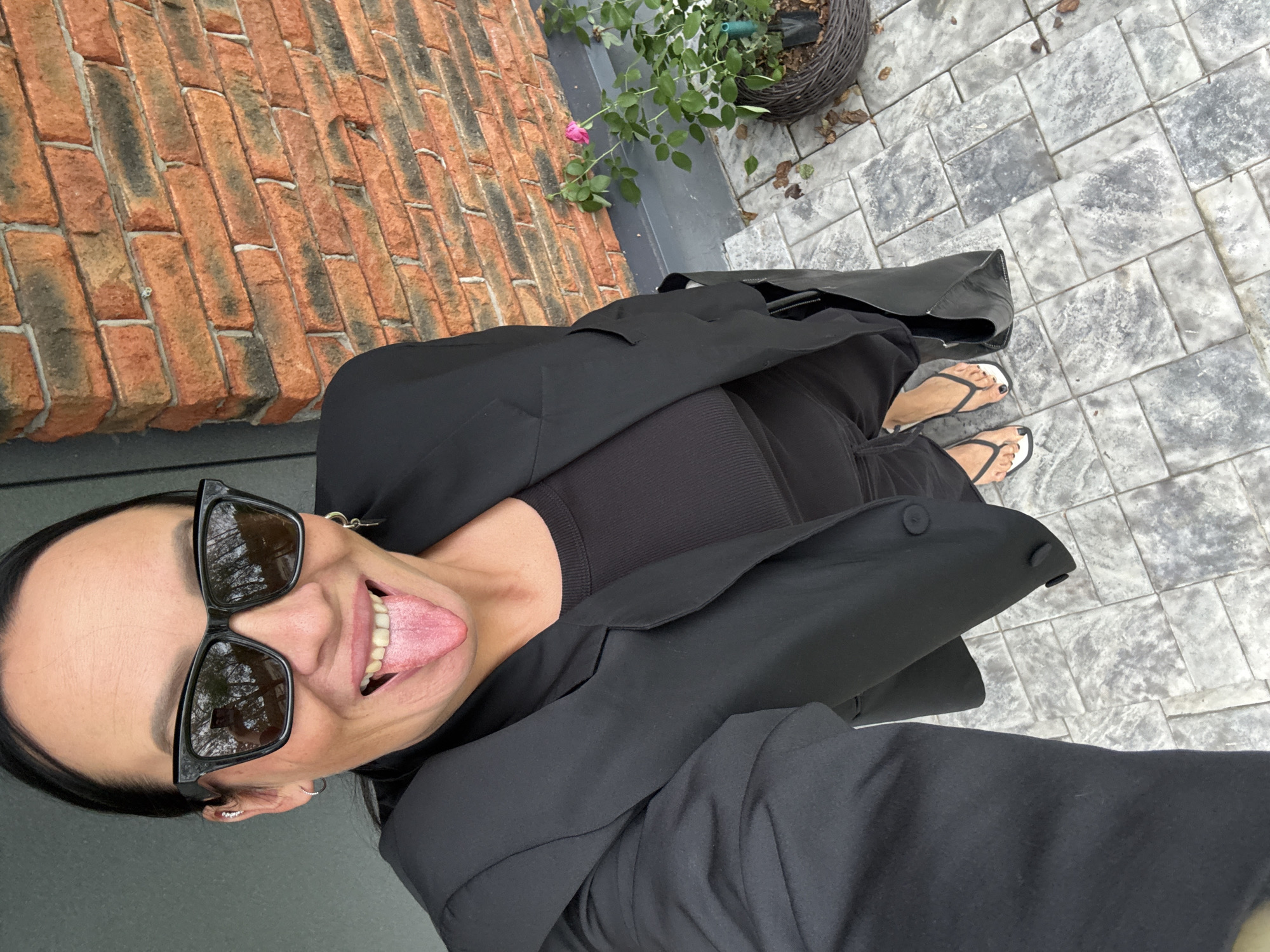A woman is taking a selfie outdoors. She is wearing a black blazer over a black top, black pants, and white flip-flops. She has on black sunglasses and is sticking out her tongue. She has pierced ears and is wearing small hoop earrings. Behind her is a brick wall and a potted rose bush with one pink flower. The ground is paved with grey interlocking stones. The lighting suggests it is daytime, possibly overcast. The scene appears to be in a residential or outdoor public area. There is no visual information in the image to confirm or deny a location in Chaikovsky, Russia.