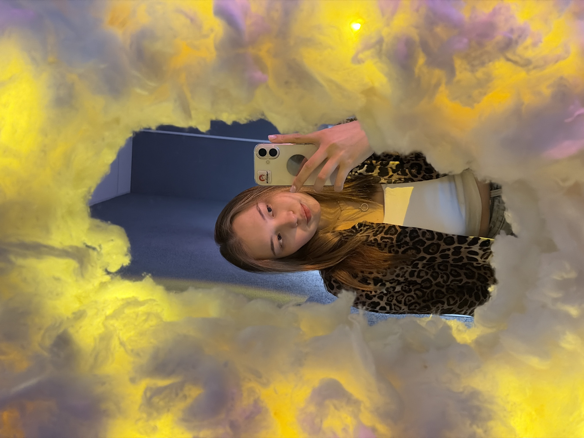 A young woman is taking a selfie in a mirror in Chaikovsky, Russia. The mirror is surrounded by fluffy, cloud-like material with yellow and pink lighting. The woman is holding a white smartphone with a clear case. She is wearing a white top under a brown and black animal print jacket. Her brown hair is down. The reflection shows a glimpse of a blue wall and gray floor.