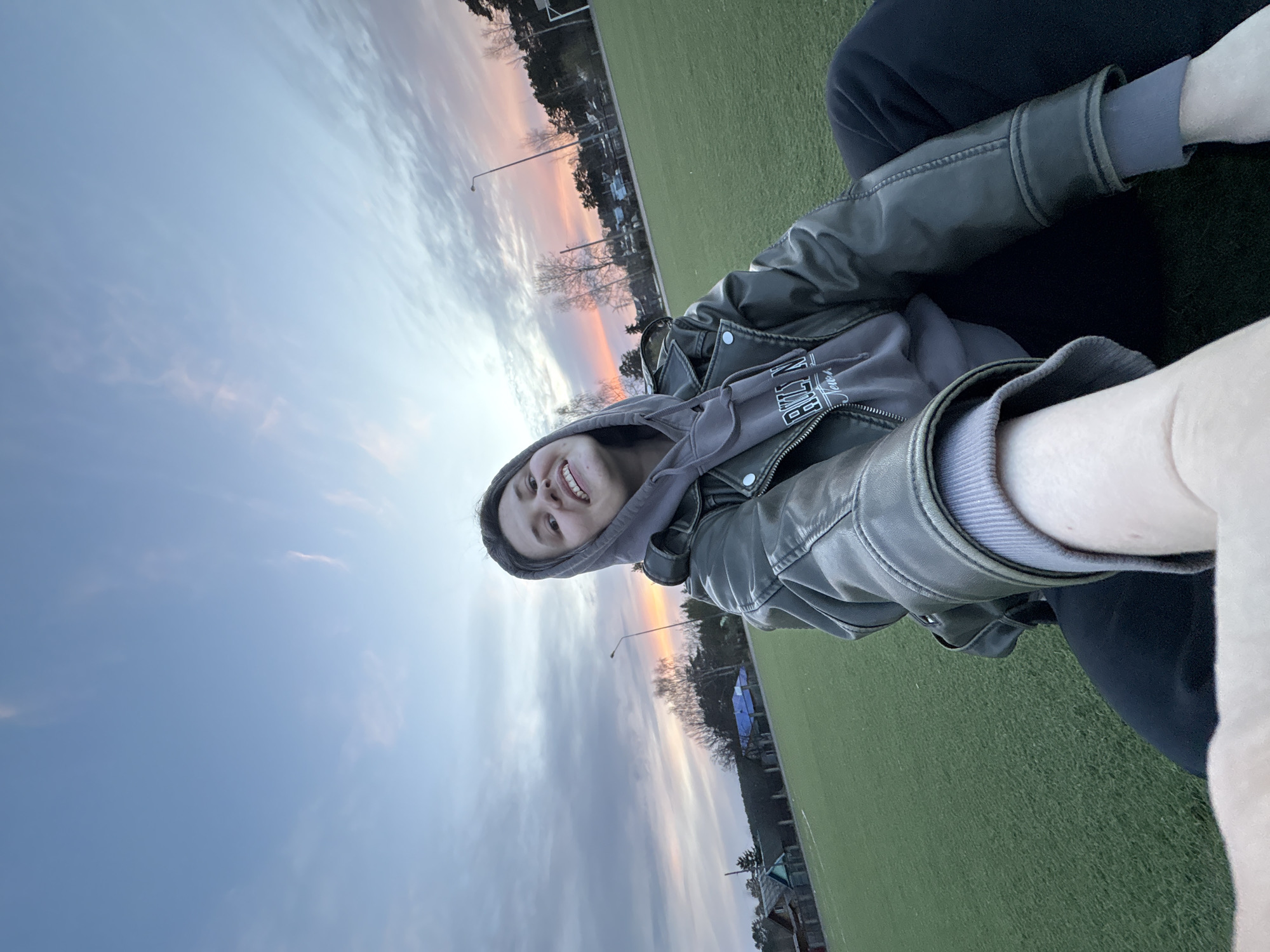 A young person is captured in a cheerful outdoor selfie, taken during what appears to be either sunset or sunrise in Chaikovsky, Russia. The individual is positioned towards the bottom left of the frame, smiling broadly and looking directly at the camera. They are wearing a grey hooded sweatshirt with the hood up, underneath a dark grey or olive-colored leather jacket. Partially visible text on the front of the hoodie appears to be "BROOKLYN." Dark pants complete their casual attire.
The setting is an open area, likely a park or sports field, characterized by a vast expanse of bright green artificial turf. This grassy area fills a significant portion of the image, especially to the right and bottom. The background showcases a scenic sky painted with soft pastel hues of blue, grey, and warm orange-pink, indicating the twilight hours. Several wispy clouds are visible. A row of bare trees stands in the mid-ground, suggesting a cooler time of year, with a prominent street light pole rising among them. Distant structures can be vaguely seen beyond the trees. The image conveys a serene and happy moment in an outdoor environment.