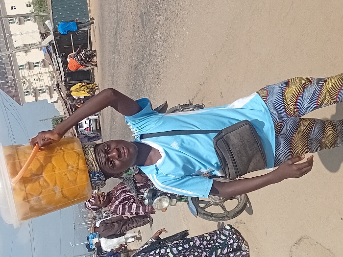 A man in a light blue shirt and patterned pants balances a large container of what appears to be food on his head. He is also wearing a dark crossbody bag. He stands next to a motorcycle on an asphalt surface, possibly a road. In the background, there are buildings, other people, and various objects that suggest a bustling outdoor setting. The image is slightly rotated, giving a tilted perspective.