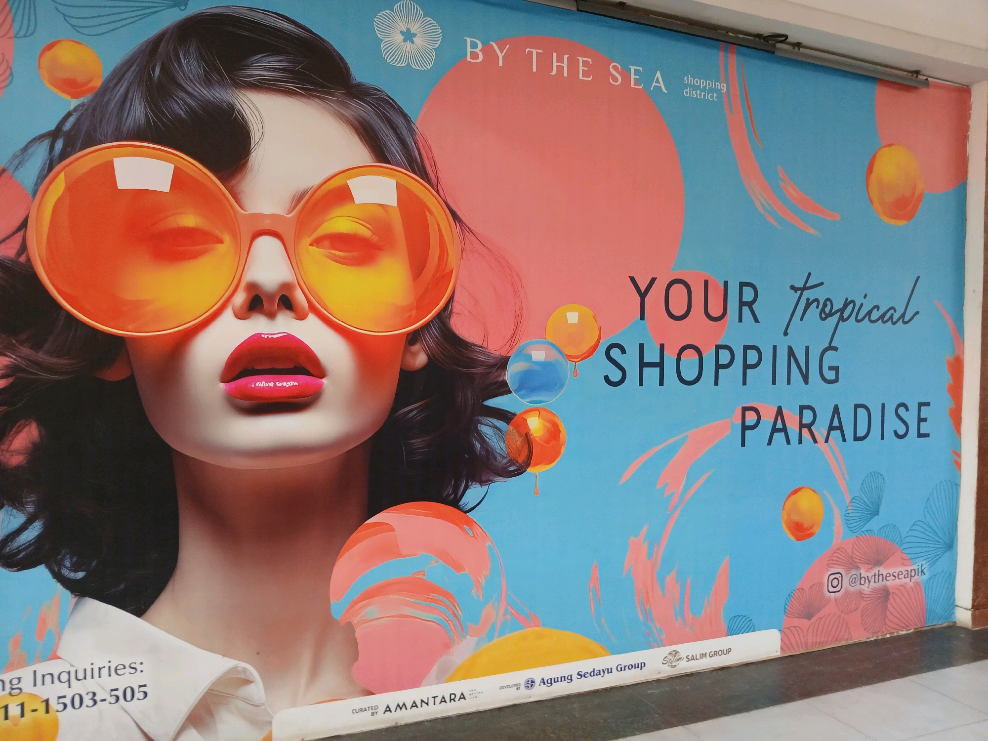 A large, colorful mural is displayed on a wall, likely in a public space. The mural features a stylized portrait of a woman with dark hair and bright red lips, wearing oversized orange sunglasses. The background is a vibrant blue with abstract elements in shades of pink, orange, and yellow, resembling bubbles or sunbursts. Text overlays the mural, with "BY THE SEA shopping district" prominently displayed at the top, and "YOUR tropical SHOPPING PARADISE" in larger font below. There is also an Instagram handle, "@bytheseapik," and several logos and text at the bottom, including "AMANTARA," "Agung Sedayu Group," and "SALIM GROUP." Based on the presence of "Agung Sedayu Group," this mural is likely located at PIK Avenue Mall in the Pantai Indah Kapuk (PIK) area of North Jakarta, Indonesia. The overall impression is of a trendy and inviting advertisement for a shopping destination.