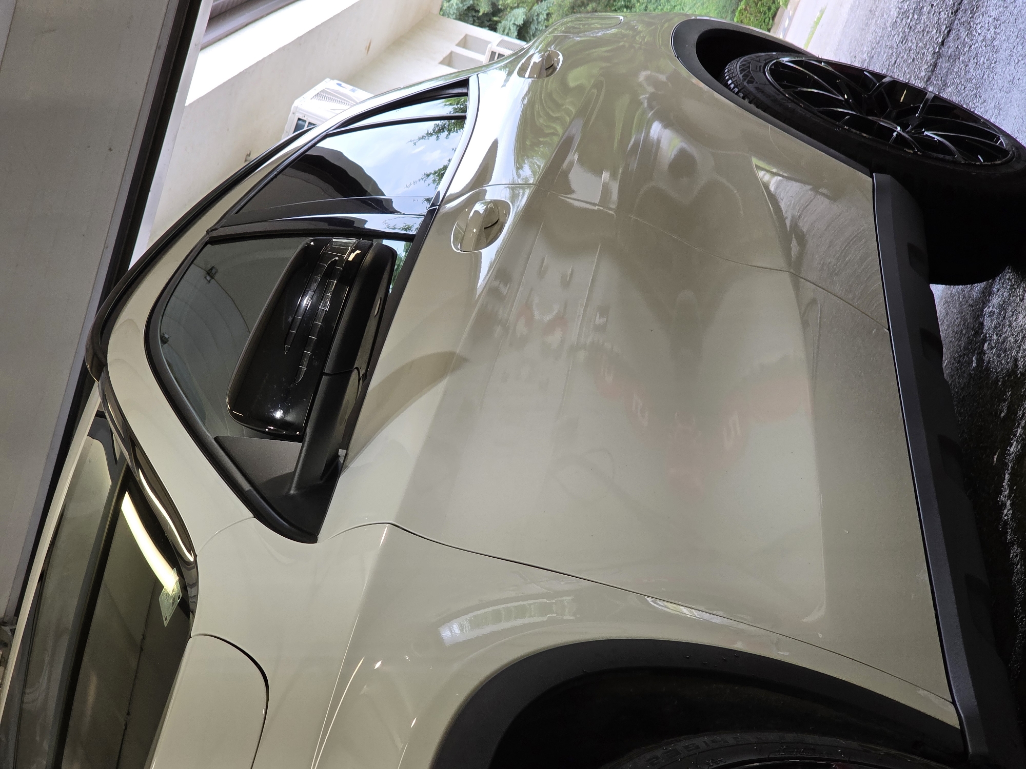 The image shows a close-up, low-angle shot of a light-colored car, possibly cream or beige, parked in what appears to be a garage or covered outdoor area. The car's body is highly reflective, showing distortions of the surroundings. The front passenger side of the vehicle is visible, with a black side mirror and tinted windows. The car has black trim around the wheel wells and distinctive black alloy wheels. The ground beneath the car is wet and reflective. In the background, through the open garage or cover, trees and a building with balconies are visible, suggesting an outdoor setting adjacent to a residential or commercial structure. The overall lighting suggests it is daytime. There are no people or any actions depicted in the image. The image does not contain any specific landmarks or features that would definitively place it in Kladnitsa, Bulgaria, or any other specific city.