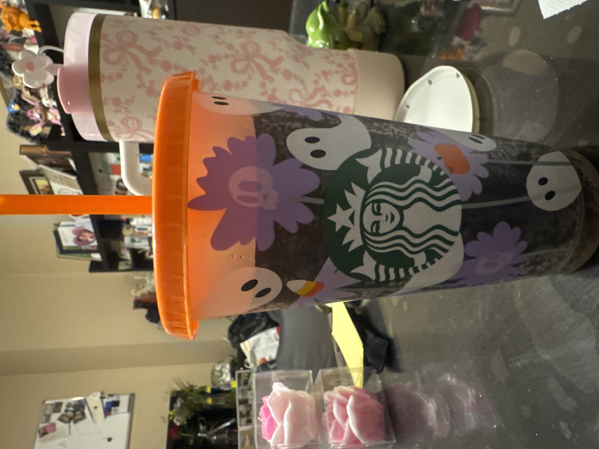 The image shows two Starbucks cups on a reflective surface. The cup in the foreground has an orange lid and is decorated with ghosts, purple flowers with skull centers, and a Starbucks logo. The cup in the background is white with pink bow designs and a flower attached to the side. The environment includes a wall, a refrigerator, and a glass surface. The objects on the desk include two small boxes containing pink roses and a yellow straw. The image location is not discernible.