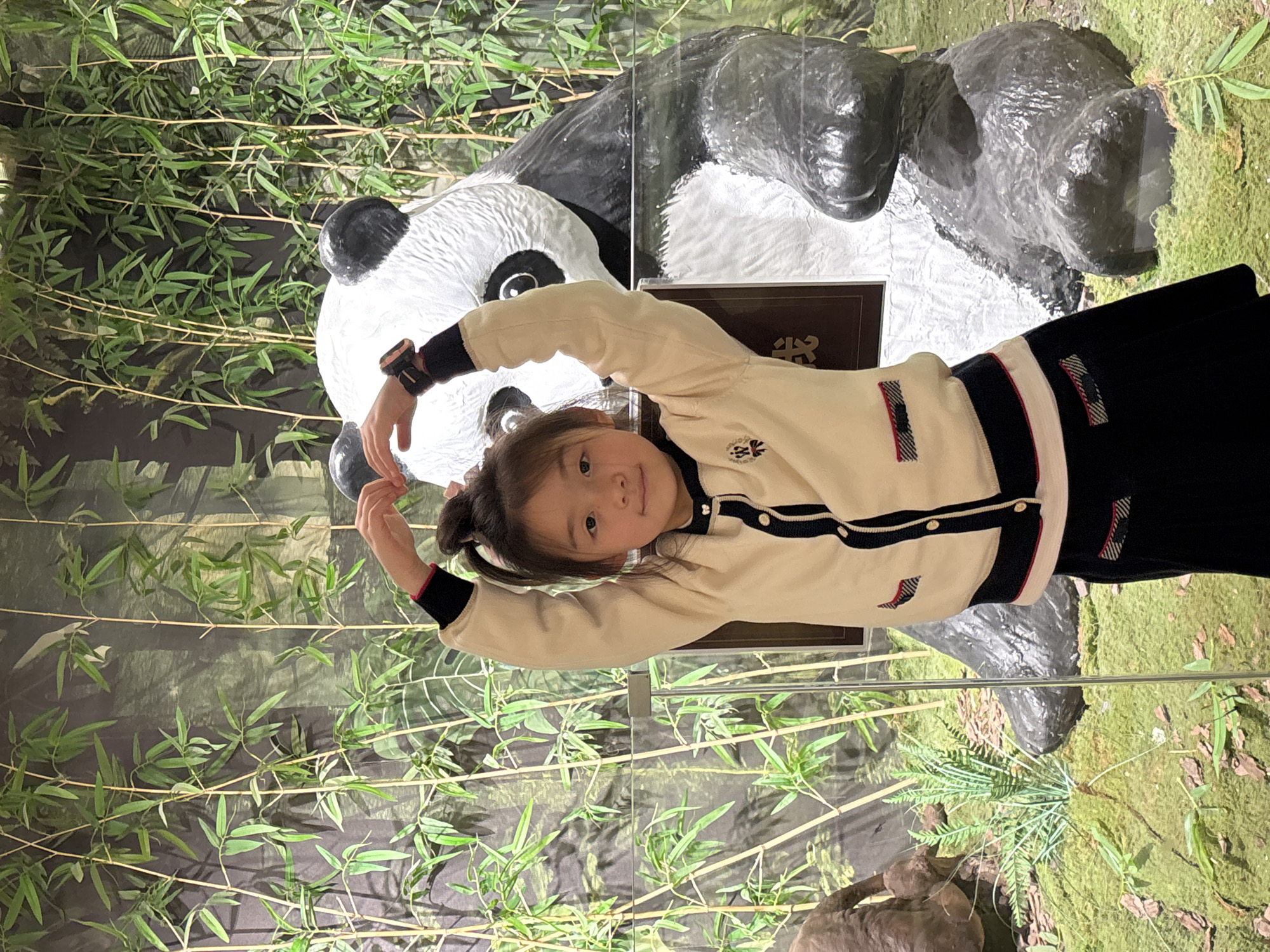 A young girl stands facing the camera, in front of a panda enclosure. The panda is either stuffed or a statue. The backdrop features bamboo. The location is likely in a zoo or wildlife exhibit in the city of Yuzhong District, China.