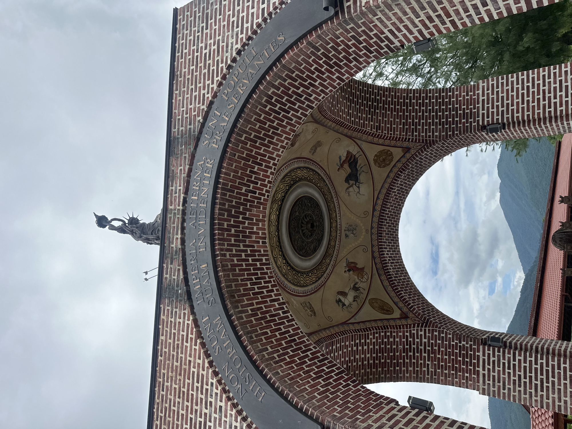 The image shows a close-up of a brick archway, likely part of a monument or building, with a statue of a winged figure holding a torch, resembling the Statue of Liberty, visible on the left. The archway is decorated with a mural on its inner surface, featuring various figures and patterns, including what appears to be a chariot pulled by horses.

Above the arch, text is inscribed on a dark metal band. The visible portion reads: "IMPERIA AE- TERNA, SUNT POPULI SUAM INVIDE NT PRAESERVANTES NON HISTORIAM". The mural and inscription suggest a historical or patriotic theme.

Through the archway, a cloudy sky and the slopes of a mountain range are visible, indicating an outdoor setting. The lighting suggests it is daytime, likely overcast due to the cloudy sky. The overall scene is a blend of architectural elements, artistic decoration, and the natural landscape. The orientation of the image is such that the sky appears at the top and the ground at the bottom, with the archway curving upwards.