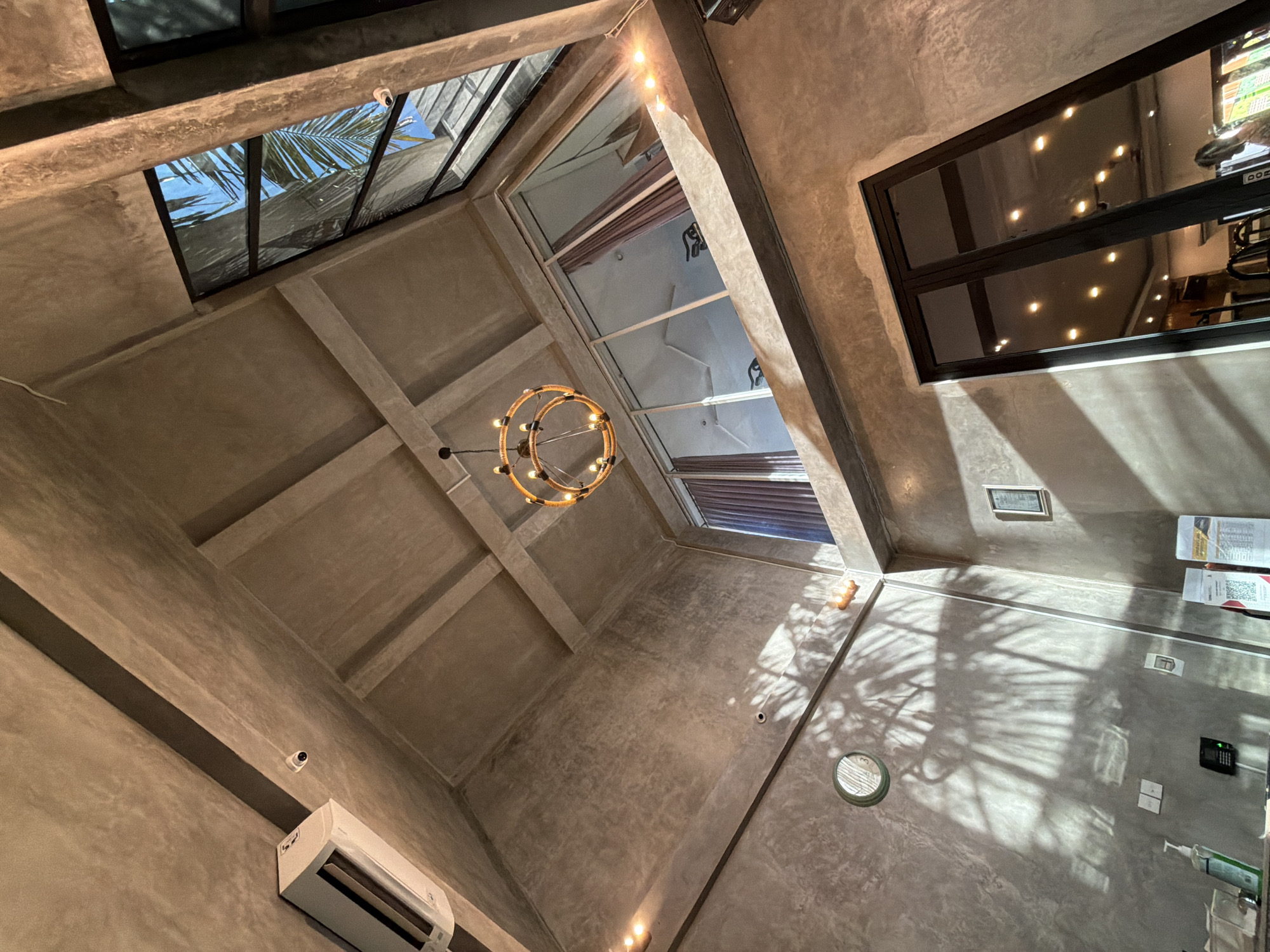 The image shows an upward-angled view of an interior space in North Kuta, Indonesia. The architectural design is characterized by exposed concrete surfaces and a network of intersecting support beams. Two skylights are visible; one on the left reveals a partial view of palm trees against a bright sky, while another directly above the frame lets in diffuse light. A circular chandelier hangs centrally in the space. Natural light casts shadows across the walls. A black-framed window is seen on the right, reflecting interior lighting. Other elements include an air conditioning unit and other wall-mounted items. There are no people or visible actions depicted in the image.