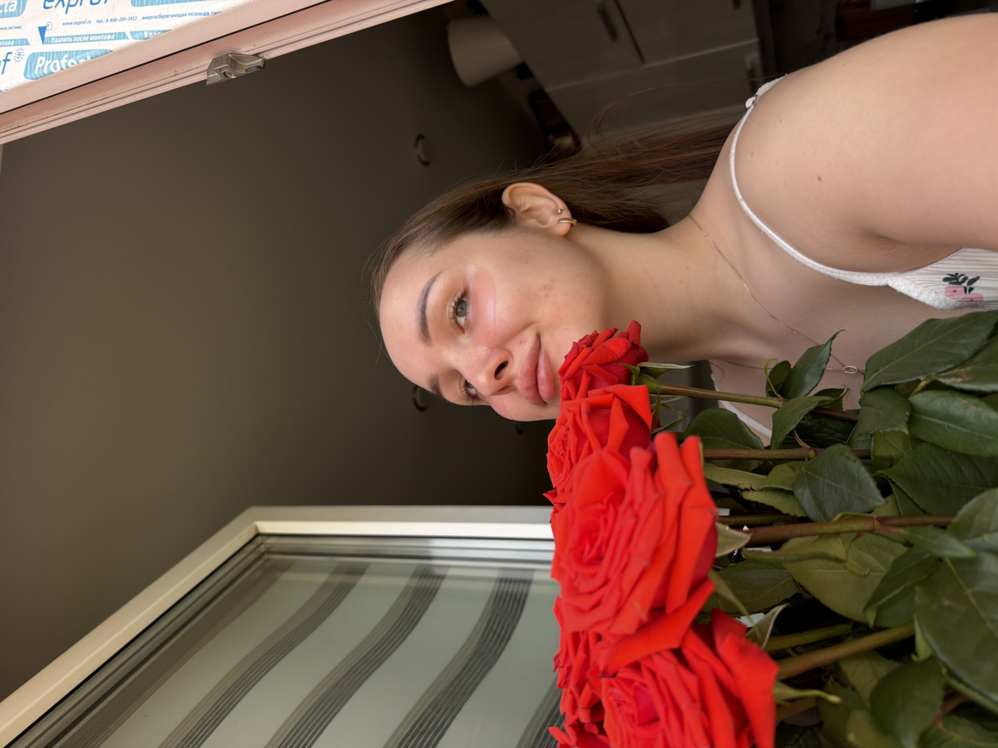 Here is a forensic description of the image:
**Scene:** An indoor setting, likely a residence. The presence of a window frame with printed text in Russian suggests a location in Russia, possibly Chaikovsky, based on the associated web address in the image.
**People:** A woman is present, positioned near the window.
**Subjects:** Red roses are prominently featured, held in front of the woman.
**Objects:** A window frame is visible, featuring text in Russian. The woman is wearing a white top with thin straps and a gold necklace.
**Actions:** The woman is holding the roses.
**Interactions:** The woman is interacting with the roses, holding them up near her face.
**Environment:** The indoor environment appears to be well-lit. A blurred interior background is visible.