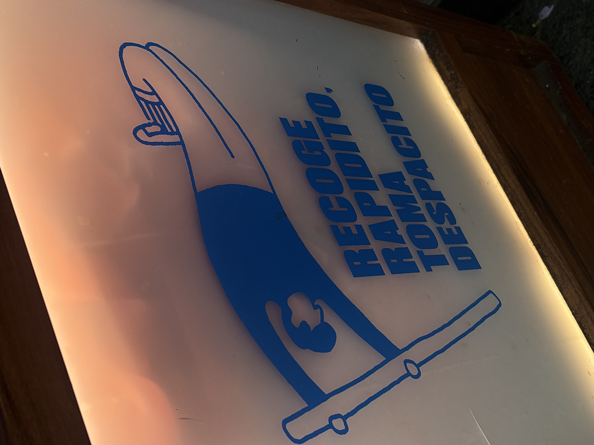 A backlit sign in Mexico City, Mexico, depicts a gymnast executing a handstand on a parallel bar. The image is rendered in blue ink on a milky white background with a wooden frame. To the right of the gymnast, text reads vertically: "RECOGE RAPIDITO, RAMA TOMA DESPACITO." The sign appears to be mounted on a surface. The photo is taken at an angle, showing perspective.