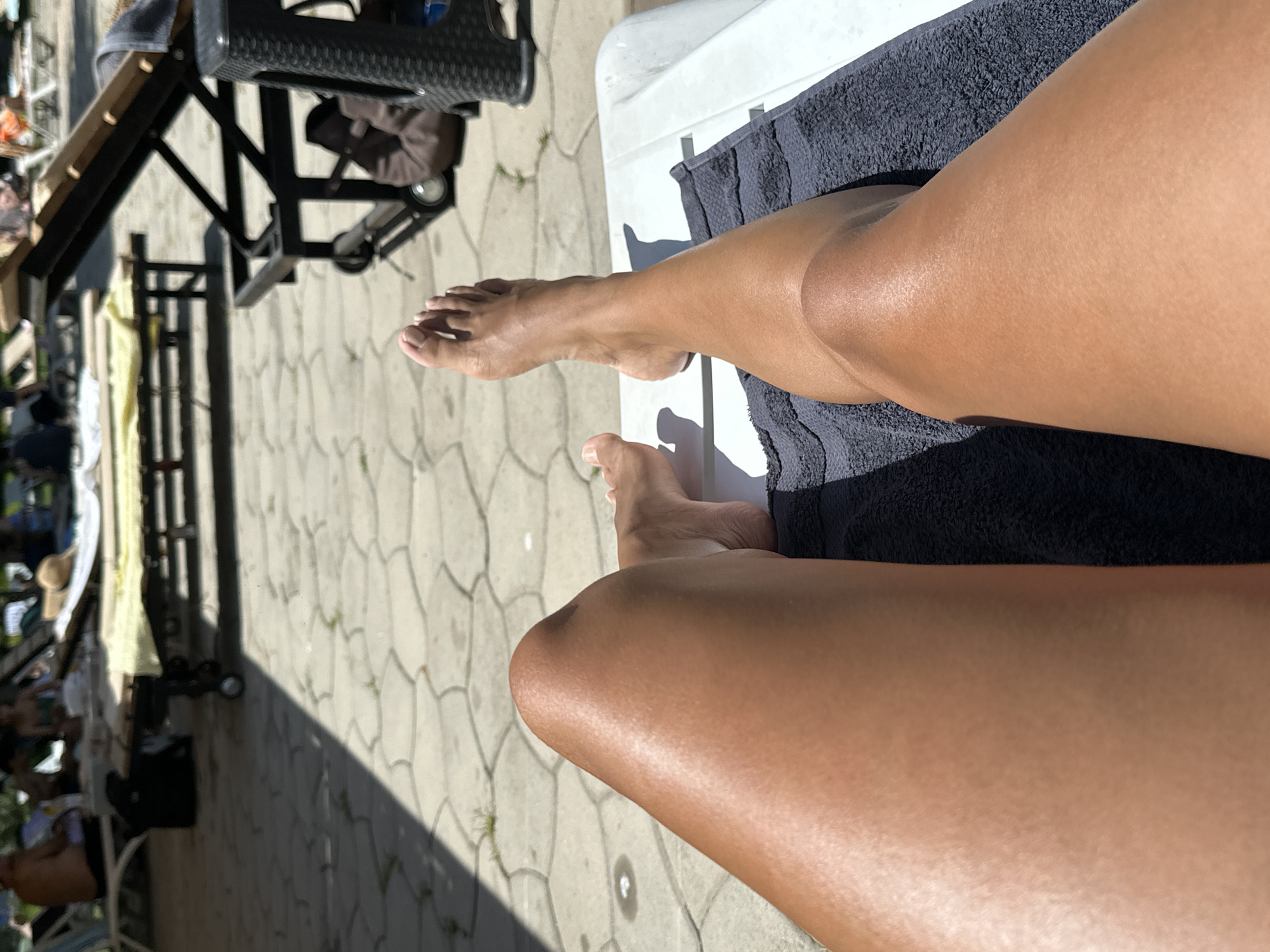The image shows a first-person perspective of a person's legs resting on a lounge chair by a swimming pool. The person's tanned legs are the primary focus, with one foot visible in the lower right portion of the frame. The background shows a paved area with a pattern of interlocking concrete stones, indicative of an outdoor public space. Several lounge chairs and umbrellas are visible, along with other people in the distance, suggesting a recreational or resort-like setting. Sunlight casts shadows across the scene, highlighting the texture of the paving stones and the sheen on the person's skin. The scene is typical of a sunny day at a public pool or resort area, and it is not possible to determine its specific location within Yerevan, Armenia, or any city.