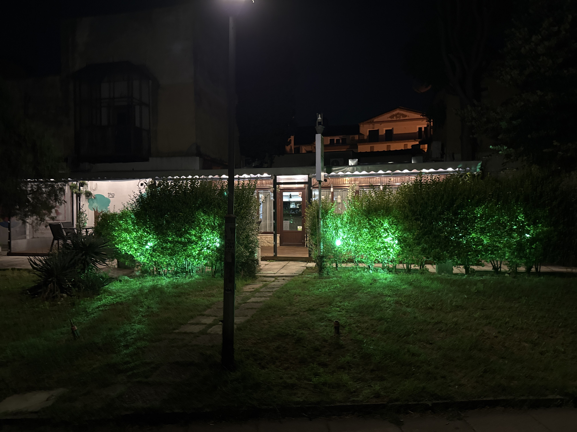 The image depicts a nighttime scene in Varna, Bulgaria, in front of a building with a covered patio area. The patio is illuminated by warm interior lights and bordered by hedges that are brightly lit with green spotlights, creating a vibrant contrast with the dark surroundings. A stone pathway leads from the foreground, across a grassy area, towards the entrance of the building. A tall lamppost stands on the left side of the path, casting some light on the scene. In the background, a multi-story building with ornate architecture is visible, with some windows illuminated. The overall impression is that of an outdoor dining or gathering space, possibly a restaurant or cafe, that is attractively lit for evening patrons. There are no people visible in the image.