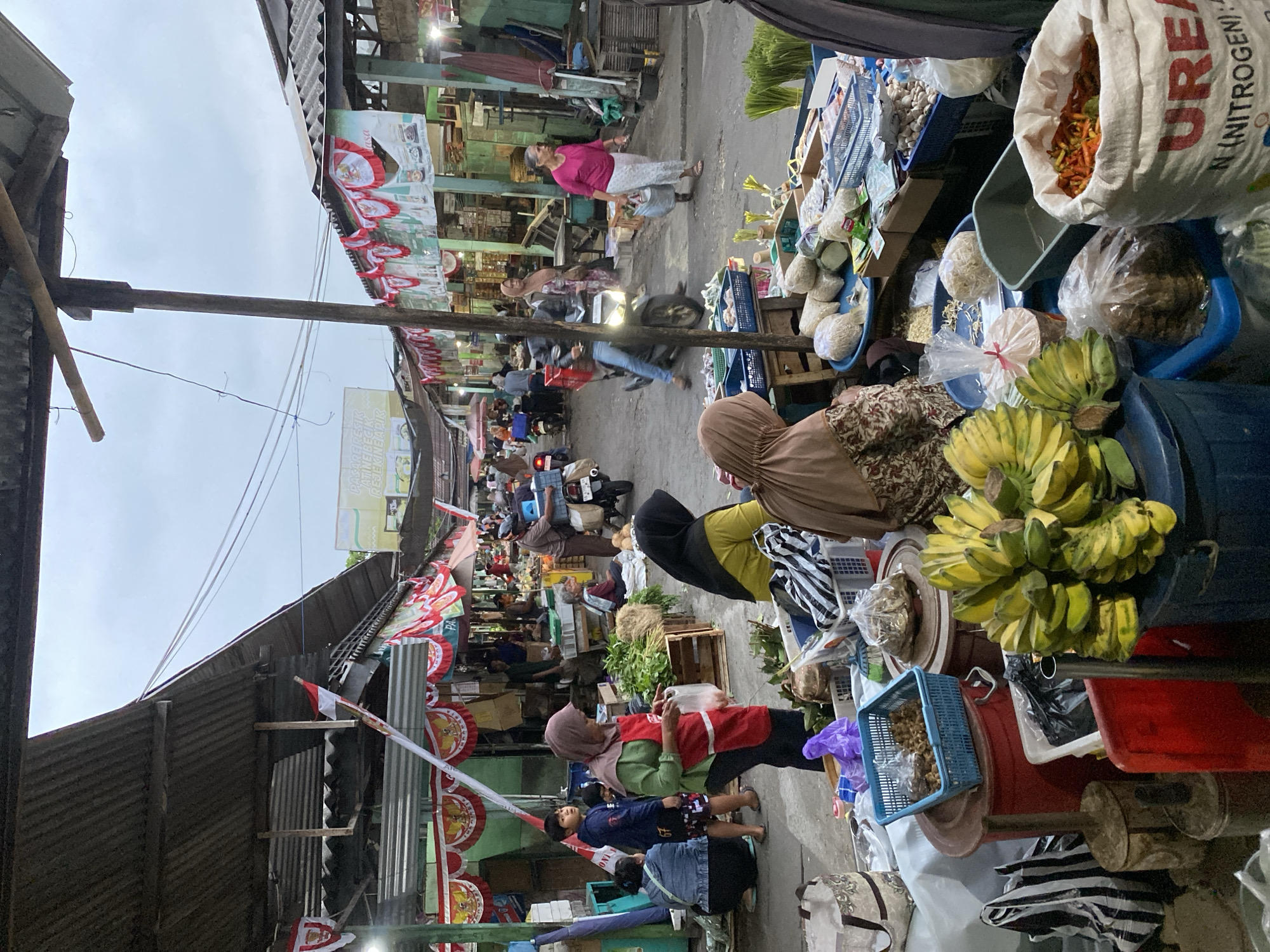 This is a street-level view of a bustling market in Ambarketawang, Indonesia. The scene is filled with stalls and vendors selling various goods, likely agricultural produce and other local wares. The market appears to be covered by corrugated metal roofs supported by wooden beams, with red, white, and blue decorative banners strung along the awnings, possibly indicating a celebration or national holiday.
Several people are visible engaging in various activities:
* **Vendors:** A woman in a red apron and headscarf is handing a plastic bag to a customer. Another woman in a yellow headscarf and dark top is seated, possibly attending to her stall. A third woman in a pink shirt and white pants is walking away from the camera, carrying a plastic bag. A man in a brown uniform is pushing a cart attached to a motorcycle.
* **Customers/Bystanders:** A young boy in a blue hooded sweatshirt and patterned shorts stands looking towards the market. A woman in a denim jacket and dark pants is crouched down. Other individuals are visible in the background, walking or interacting at the stalls.
* **Objects:** Prominently displayed are bunches of yellow and green bananas. Several sacks are present, one white sack with red lettering that appears to say "UREA N (NITROGEN)". Baskets filled with various items, including what look like dried goods, chilies, and produce, are visible. Blue and red plastic containers are also scattered throughout the scene. Bags of various sizes and patterns, including striped ones, are stacked or placed on the ground.
The environment is a street lined with market stalls. The ground is paved. The sky is overcast, suggesting it might be morning or late afternoon. The overall atmosphere is one of lively commerce and community activity. The presence of the decorative banners suggests a specific occasion or cultural context within Ambarketawang.