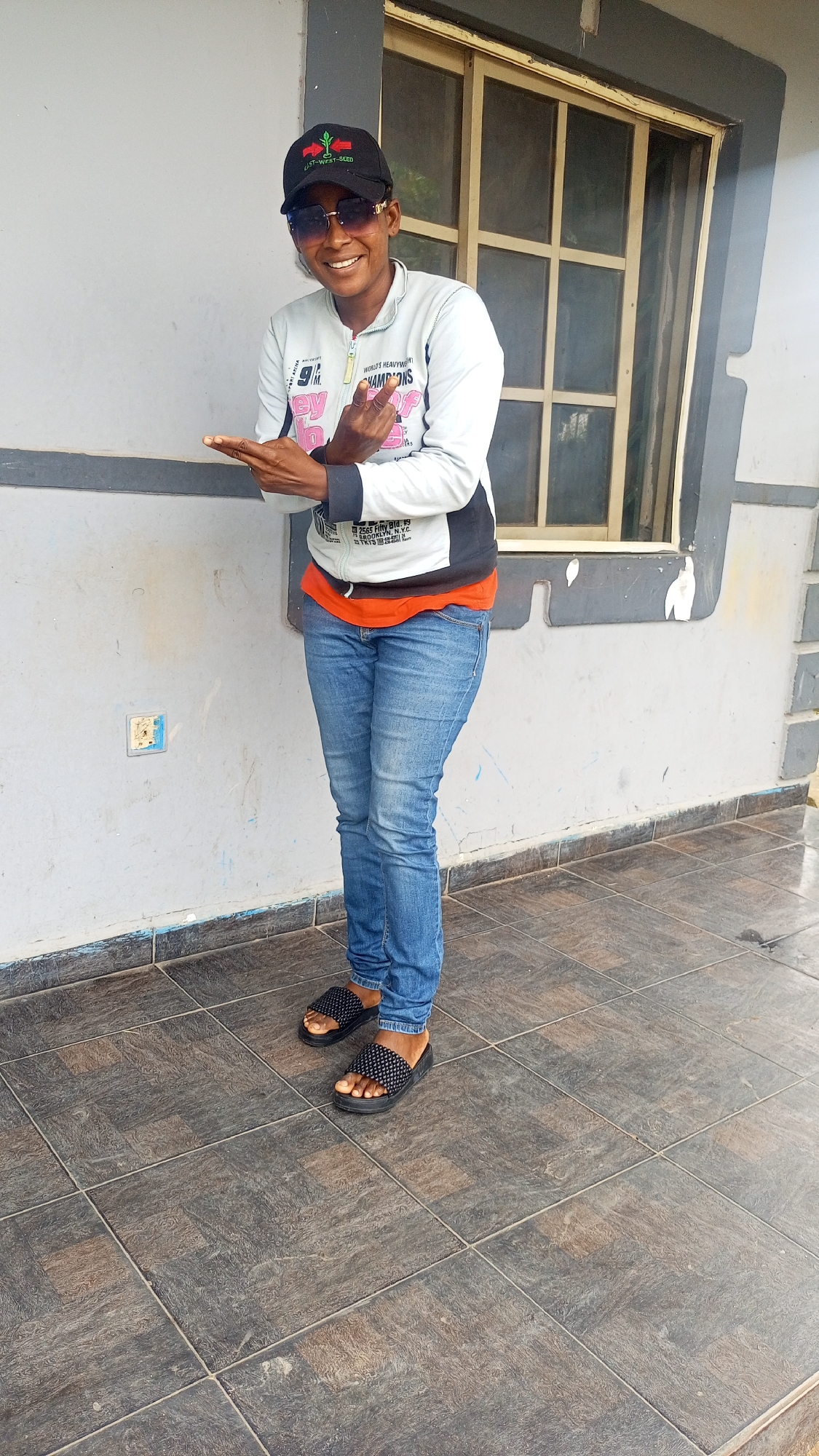 A young Black female wearing a black baseball cap with a red and green logo, sunglasses, a light gray zip-up hoodie over an orange t-shirt, and blue denim jeans stands on a tiled walkway in front of a light gray building. She is smiling and facing the camera, with her left arm extended slightly and her right hand in a peace sign gesture. The building has a window with a grid pattern, and there is a light switch on the wall to the left of the subject. The tiled floor is a mosaic of dark gray and brown squares. The overall scene appears to be an outdoor setting in a residential area. There is no specific indication that the location is Rido, Nigeria.