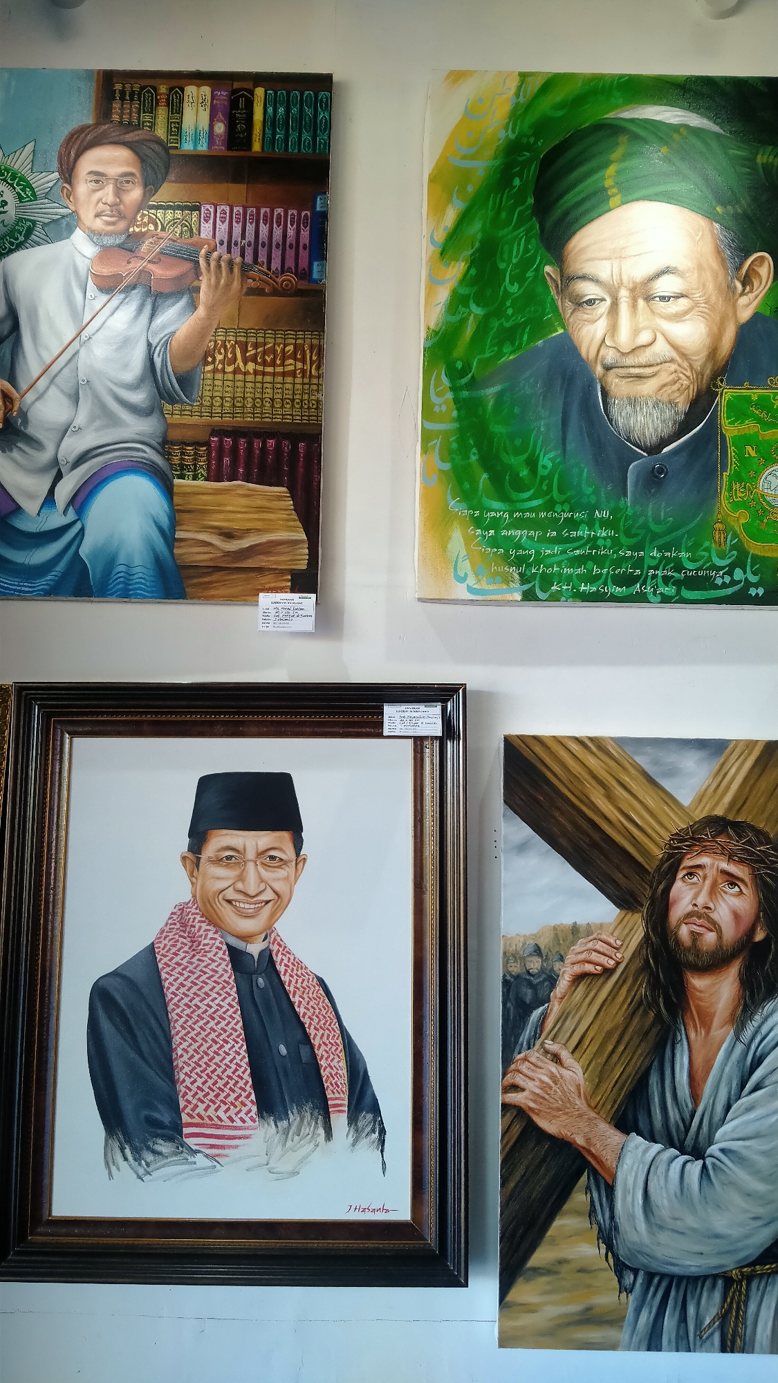 The image displays a collection of four paintings on a white wall, possibly in a gallery or exhibition space. The paintings are arranged in two rows, with two paintings in the top row and two in the bottom row, though the bottom row is partially obscured.
The top left painting depicts an elderly man wearing a turban and traditional clothing, playing a violin. He is positioned in front of a bookshelf filled with books.
The top right painting is a close-up portrait of an elderly man with a green turban and a beard, looking downwards. The background features green calligraphy.
The bottom left painting is a framed portrait of a smiling man wearing a black cap, a dark suit, and a red and white patterned scarf. His name, "J. Hszanz," is visible in the bottom right corner of the painting.
The bottom right painting shows a representation of Jesus Christ carrying the cross. He is depicted with a crown of thorns and is looking upwards, with a crowd of figures in the background.
A small white label is attached to the wall below the top left painting, and another label is attached to the frame of the bottom left painting. There is no visible indication of the location being the city of Nyamat, Indonesia.