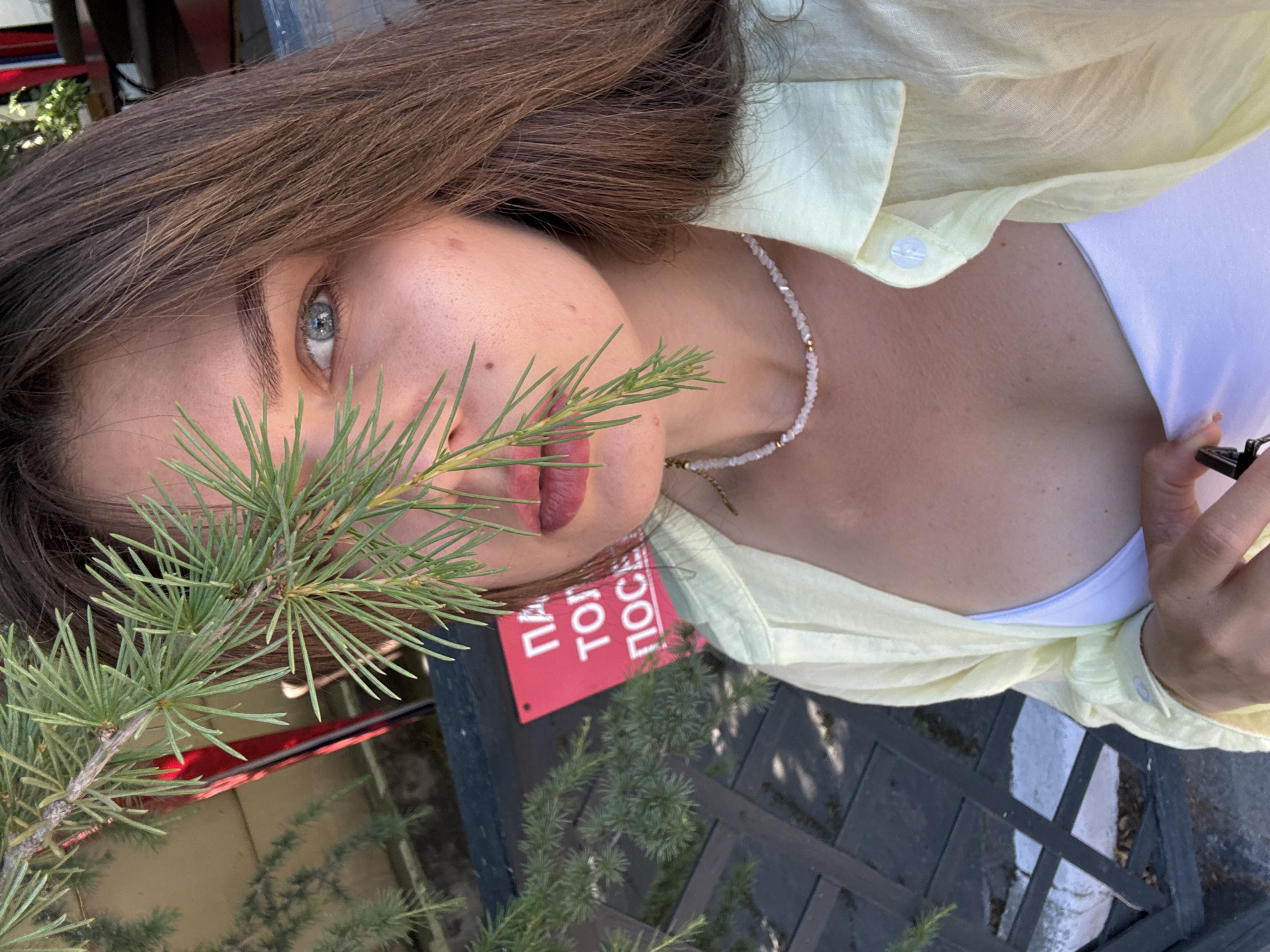 The image shows a close-up, head-and-shoulders shot of a young woman with brown hair and blue eyes. She is wearing a light yellow collared shirt over a white top and a beaded necklace. Part of a pine branch is in the foreground, obscuring her face slightly. Behind her, a red sign with white Cyrillic text is visible. The text on the sign reads "ПАЛ ТОЛ ПОСЕ," which is likely part of a larger phrase. The setting appears to be outdoors, with wooden surfaces and foliage visible in the background. The lighting suggests it is daytime. The woman's expression is neutral.