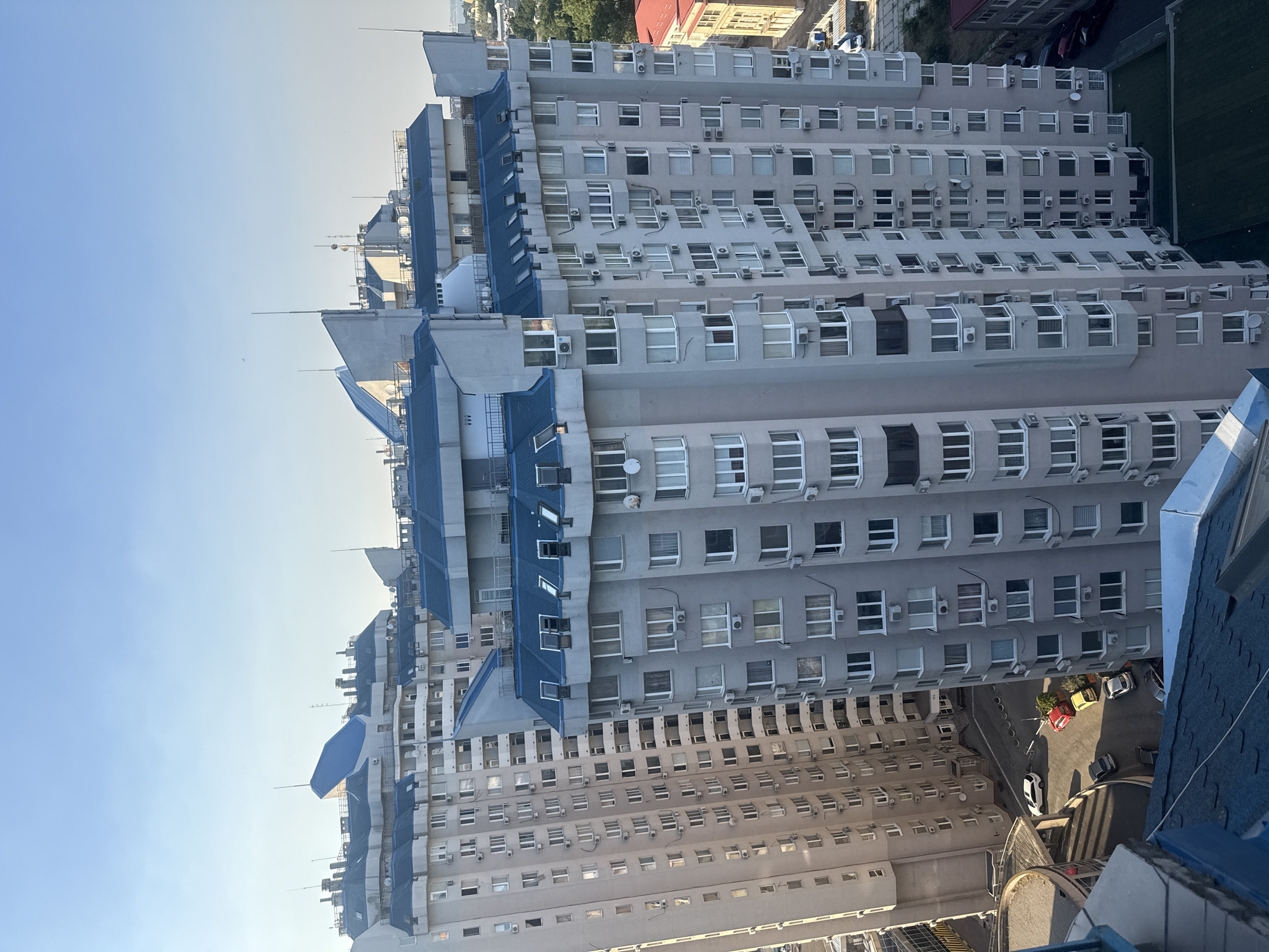 This media file captures an aerial view of a series of interconnected, multi-story buildings with a distinctive light gray facade and blue-tiled roofs. The buildings appear to be residential or office complexes, with numerous windows and individual air conditioning units visible on their exteriors. The architectural style suggests a modern urban setting.
The scene is bathed in natural light, indicating it is daytime, likely morning or afternoon, with a clear blue sky dominating the upper left portion of the image. The angle of the sunlight casts shadows, highlighting the varied depths and protrusions of the building structures.
In the lower right portion of the image, a street level is visible with several parked cars, including a red sedan, a yellow car, and a white SUV. A patch of green grass or a small landscaped area is also present adjacent to the street. The presence of buildings, street infrastructure, and vehicles places the scene firmly in an urban environment.
While there are no people visible in the image, the overall impression is one of a densely populated urban area. The clean lines and orderly arrangement of the buildings suggest a well-maintained and modern part of the city. The image is oriented vertically, giving a sense of looking down upon the city. No visible text is present in the image. The location context provided indicates Odesa, Ukraine.