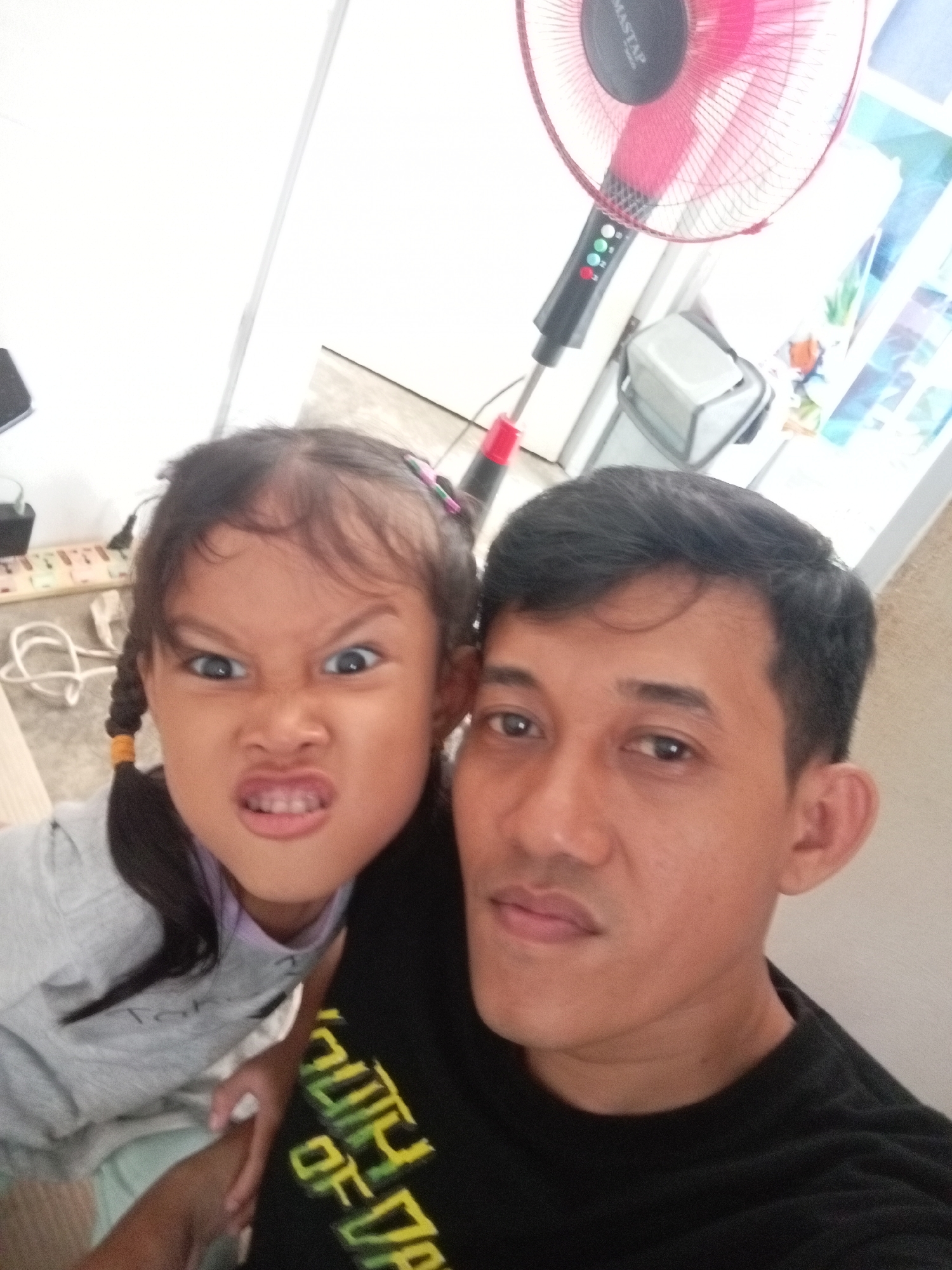 The image is a selfie taken indoors, likely at a residence in Batam, Indonesia. A man and a young girl are the primary subjects. The man, positioned on the right, has short dark hair and is wearing a black t-shirt with a graphic design. He is looking towards the camera with a neutral expression. To the left of the man, the girl is making a funny, exaggerated angry face, with her eyes wide and mouth contorted. She has dark hair styled in two braids and is wearing a light gray long-sleeved shirt. In the background, a red and black standing fan is visible, along with other household items such as a white bin and electrical cords. The overall environment appears to be a living space or common area within a home.