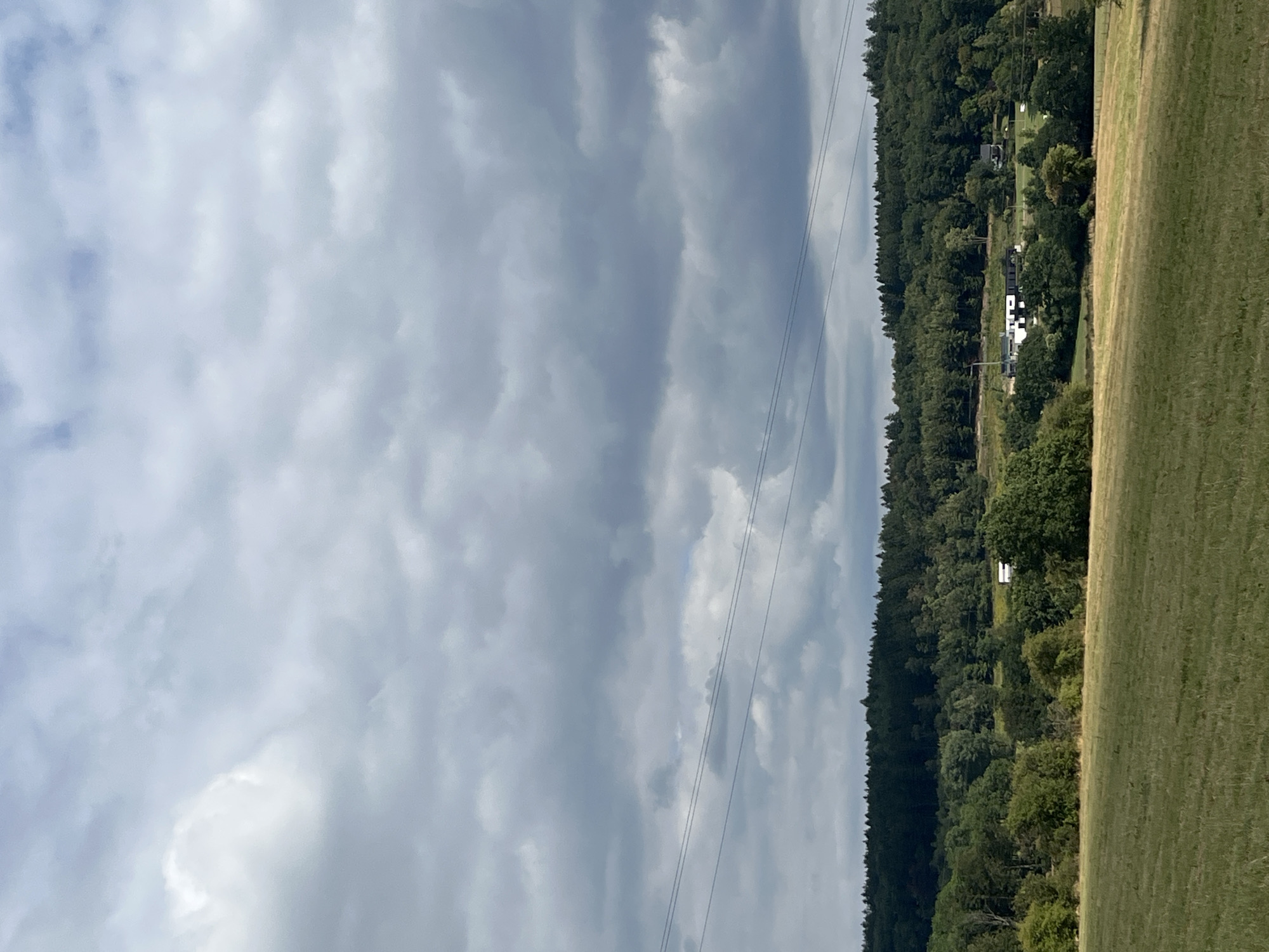 This image captures a rural landscape near Saint Vith, Belgium, characterized by a cloudy sky, rolling hills, and a dense forest. In the foreground, a mown field stretches towards a line of trees. Beyond the trees, a few buildings are visible, including a modern-style house with a black roof and white walls, and a white caravan. A line of power lines crosses the middle ground of the image, suspended above the forest. The sky is filled with dramatic, dark clouds, suggesting overcast weather. The overall scene conveys a sense of natural beauty and tranquility, with a touch of human presence in the form of the scattered structures.
