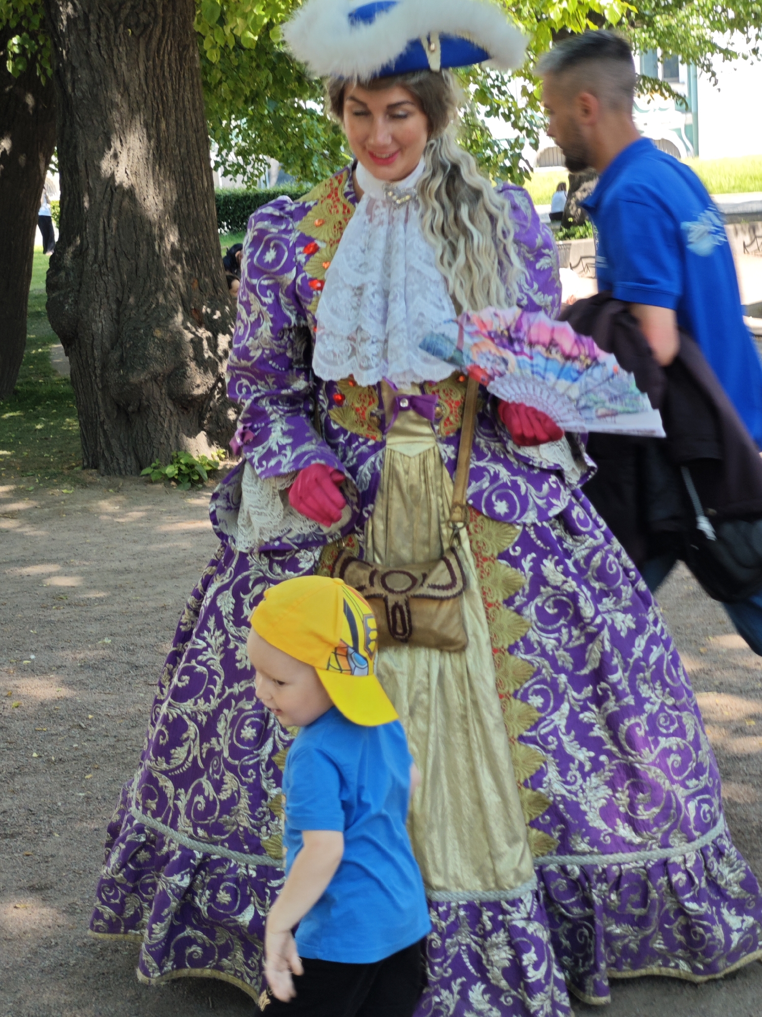 This is a medium shot photograph taken outdoors, featuring a woman dressed in an elaborate historical costume and a young child. The woman is wearing a voluminous purple gown with gold ornate patterns, a ruffled white lace collar, a feathered blue hat, and red gloves. She is holding a decorative fan with colorful patterns. A brown leather pouch is slung across her body. The child, positioned in the foreground and to the left of the woman, is wearing a bright yellow baseball cap and a blue t-shirt. The child is looking away from the camera and appears to be in motion, with their right hand extended.
In the background, a man in a blue polo shirt is walking away from the camera. Further back, another person is visible in the distance. The environment suggests a park or outdoor event setting, with large trees, gravel pathways, and some greenery. The overall atmosphere appears to be casual and possibly festive. While the image does not explicitly state the location, the architecture visible in the background, particularly the building with green trim, is consistent with the style found in St. Petersburg, Russia. However, without more specific visual cues, this remains an assumption based on stylistic similarities. The lighting suggests a bright, sunny day.