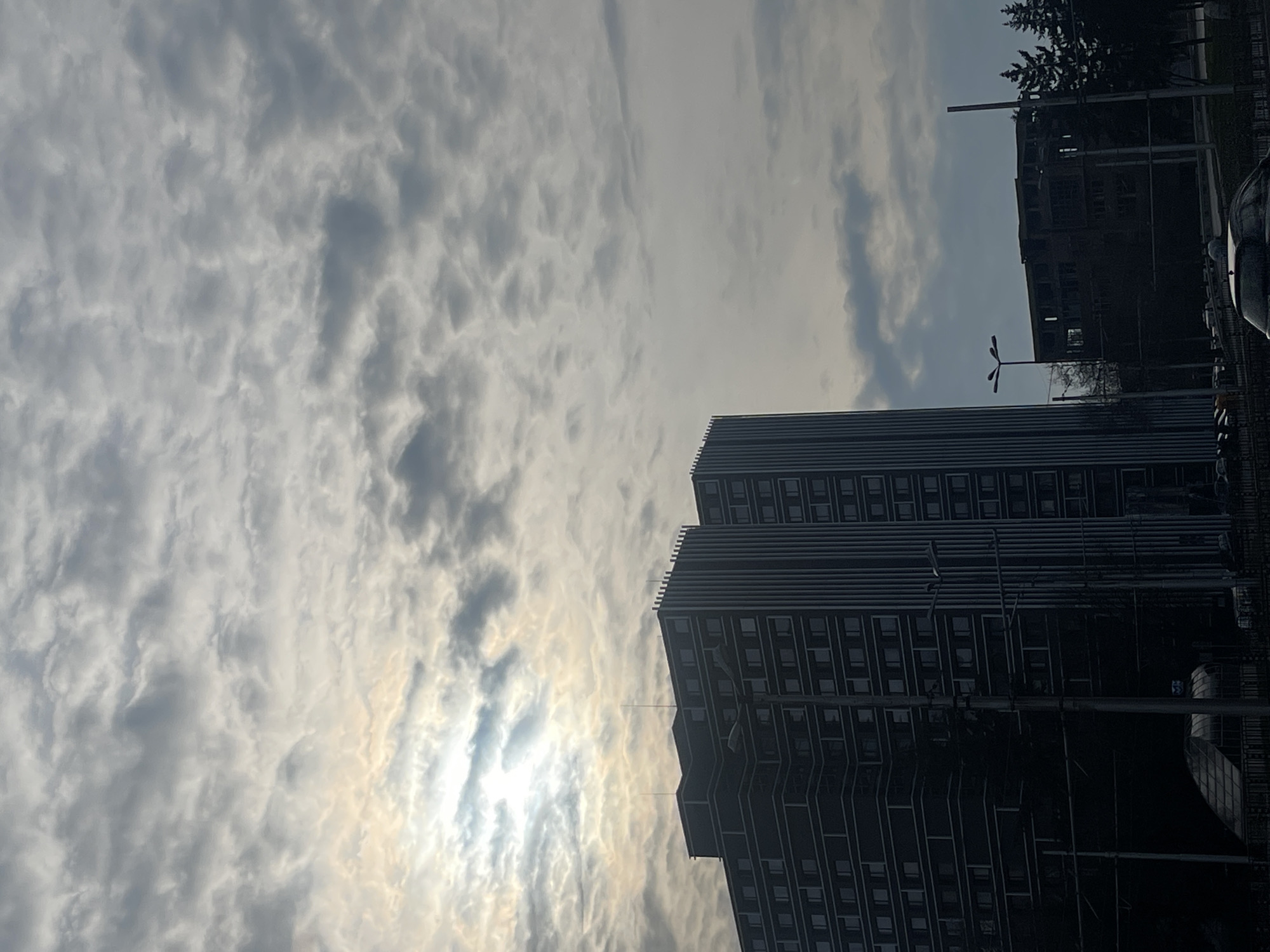 The image depicts an urban scene in Sofia, Bulgaria. The dominant element is a tall, modern building with a dark facade and numerous windows, possibly an apartment complex or office building. The building appears to have a stepped design with multiple sections of varying heights. To the right of this structure is another building that appears to be under construction or partially damaged. The sky is filled with dense clouds, with the sun faintly visible through a thin spot, creating a bright area surrounded by cloud cover. Street lamps are seen in the foreground, as well as what looks like a guardrail and a parked car. The overall composition suggests a late afternoon or early morning light, with the buildings appearing in silhouette due to the strong backlighting from the sun.
