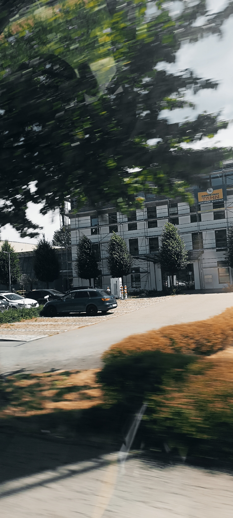 This is a motion-blurred photograph taken from a vehicle moving through Winterbach, Germany, on a sunny day. In the foreground, the blurred motion of trees and foliage suggests the viewer is looking out from behind branches. A dark gray station wagon is parked on a paved surface, with other vehicles partially visible to its left. Behind the vehicles, a modern, multi-story building is under construction, indicated by scaffolding surrounding its exterior. A sign on the building reads "TG GERÜSTBAU TAHN," identifying a scaffolding company. The building has many windows and appears to be a commercial or office space. The ground outside the building is a mix of paved areas and vegetation. The overall scene depicts a street view of a developing urban area, captured with a sense of movement.