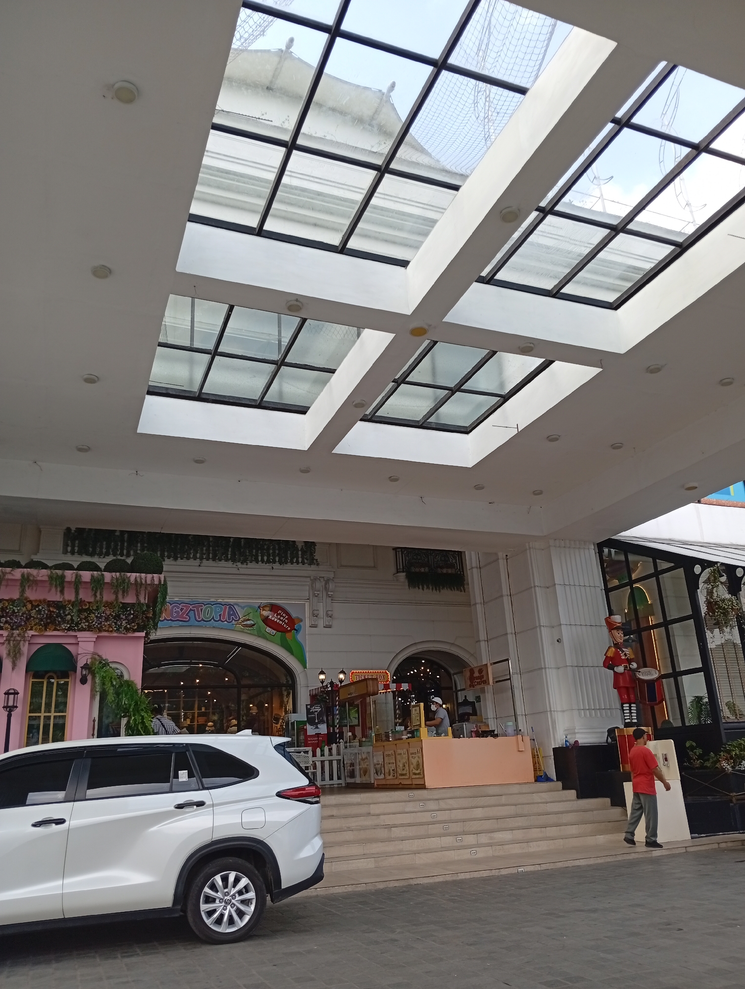 The image captures an outdoor scene in front of an entertainment venue with a large, multi-paneled skylight overhead. A white SUV is parked to the left, partially obscuring the entrance to the venue. The entrance itself is adorned with hanging greenery and features a pink facade on the left side, with a large arched entryway. Inside the entrance, there are various food stalls and a sign that reads "KINGZ TOPIA". A person in a red shirt and gray pants is walking away from the entrance towards the right side of the frame. To the right of the entrance, a life-sized nutcracker-like figure in a red uniform stands guard. The ground is paved with interlocking bricks, and there are steps leading up to the entrance. The overall impression is of a brightly lit commercial area, possibly a mall or amusement park entrance. The scene is likely located in the city of Cimacan, Indonesia, based on contextual information.