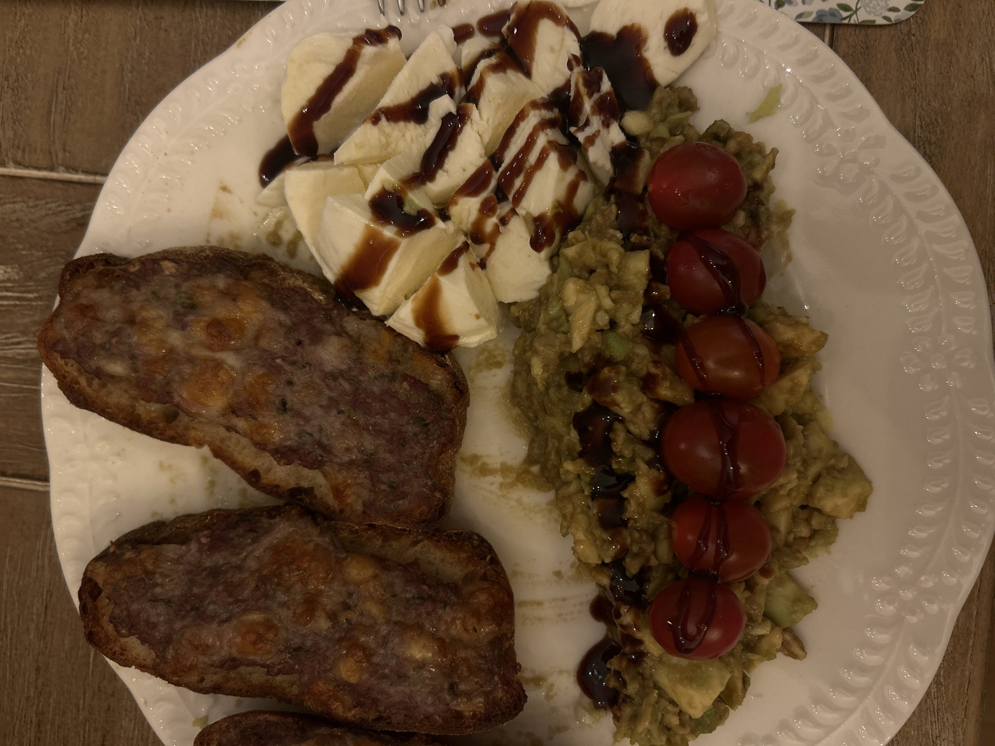 A white plate contains a meal consisting of toasted bread with a topping, mozzarella cheese drizzled with a dark sauce, mashed avocado, and a row of cherry tomatoes also drizzled with the same dark sauce. The photograph was taken in an unknown location. There is no specific indication that this is in Plovdiv, Bulgaria. No people, actions, or interactions are visible. The environment consists of a wooden surface beneath the plate.