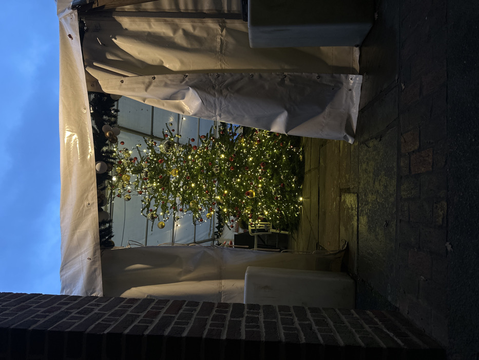 The image depicts an outdoor night or dusk scene in Sevenoaks, United Kingdom, viewed through a narrow opening. A large, conical Christmas tree stands centrally within a white, opaque tent structure. The tree is extensively adorned with numerous warm white lights, spherical red ornaments, and spherical gold ornaments. Some silver-colored baubles are also visible on the tree and as part of garlands draped from the tent's internal framework.
The perspective is framed by a dark brick wall column on the left and a partially drawn white tent flap on the right. A white, rectangular object is positioned on the ground to the left of the tent flap. The ground immediately outside the tent entrance is dark and paved with flagstones and possibly brick, showing reflective patches indicative of dampness. Inside the tent, the ground surface consists of flagstones, illuminated by the tree's lights. A dark blue, overcast sky is visible above the tent canopy. No individuals are present in the scene. Partially obscured in the lower-left background within the tent are what appear to be a white chair and a dark object, possibly a table or counter.