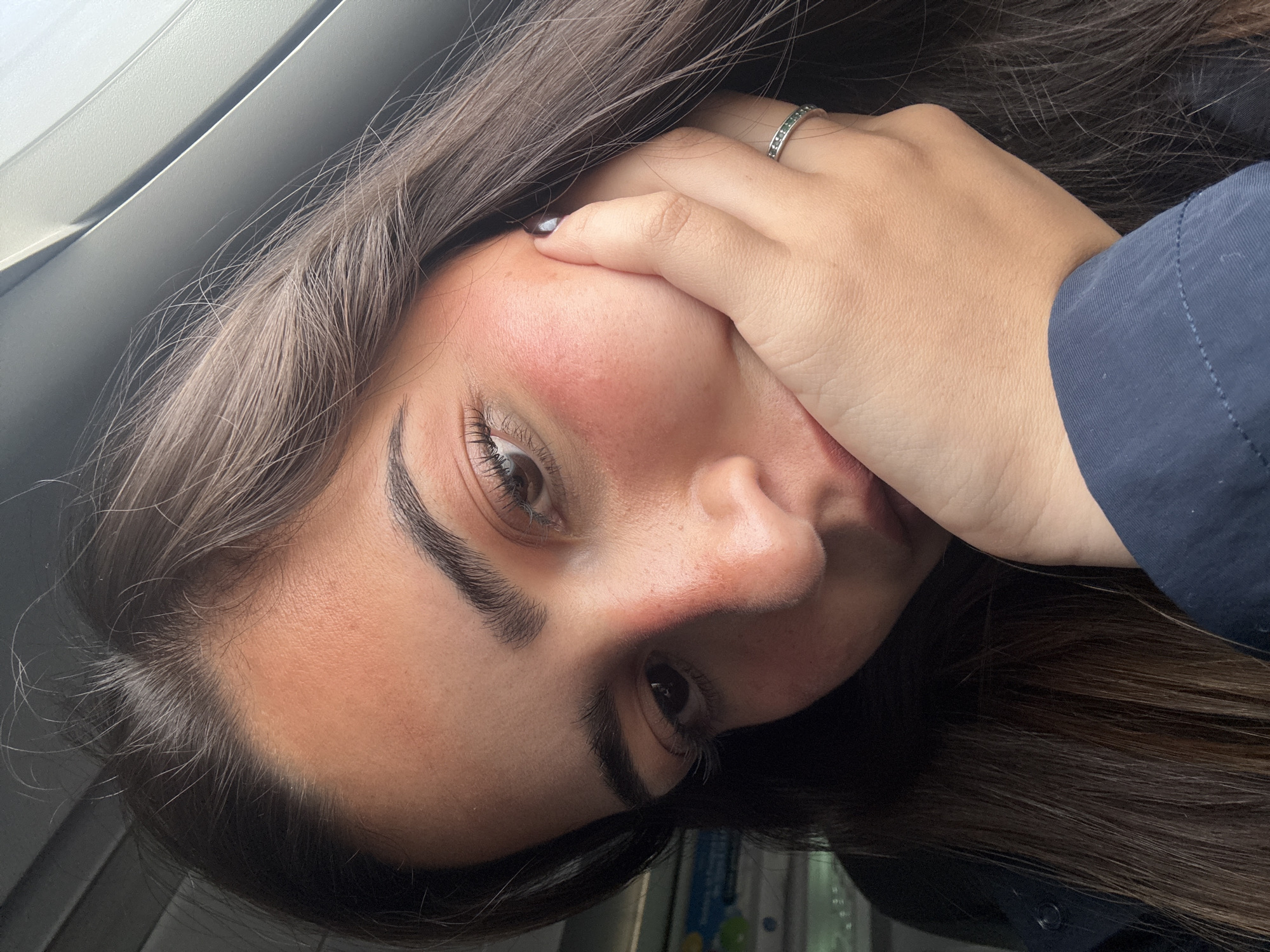 A close-up portrait shows a young woman with long, dark brown hair holding her hand to her face. She is wearing a ring on her ring finger of her left hand. Her skin is pale with reddish patches on her cheeks and nose. The background appears to be a window with grey trim and potentially a glimpse of the interior of a vehicle or building. Given the prompt's requirement of specifying a location in San Antonio, United States of America, it is impossible to determine the specific location without additional information. Therefore, the location is unspecified.