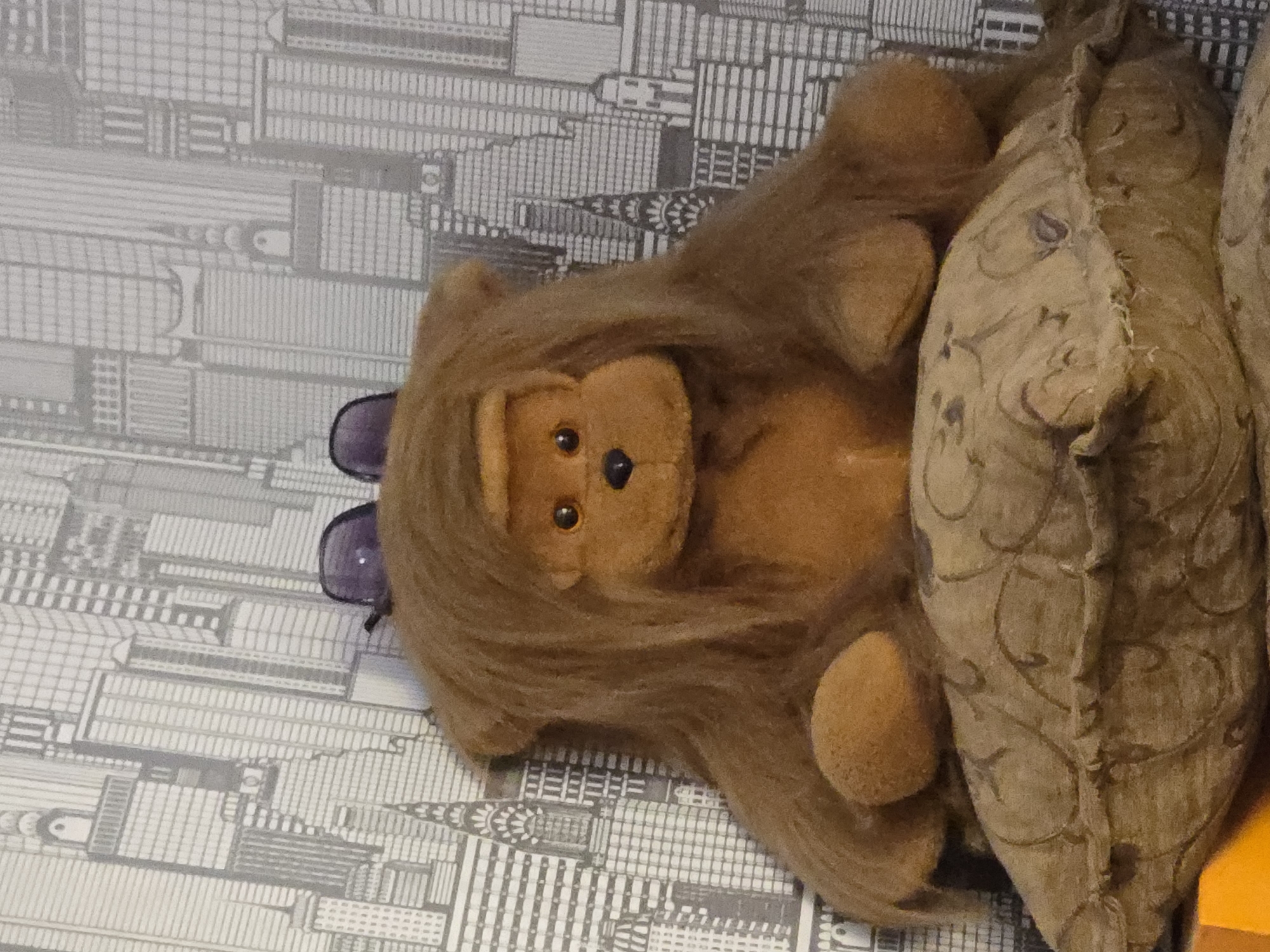 A brown, plush lion toy with a mane, a tan face with black button eyes and a black nose, and purple glasses is lying on its back against a large, patterned cushion. The lion is positioned vertically in the frame, its head towards the bottom left and its paws towards the top right. The background is a grey and white wallpaper with a pattern resembling a city skyline, featuring many tall, thin buildings. The cushion has a textured, woven appearance with an ornate, swirling pattern in a darker shade of brown. The lighting appears to be artificial, casting soft shadows on the subjects. No people are visible, and there is no indication of a specific time of day or weather. The scene appears to be indoors, possibly in a room. No text is visible in the image.