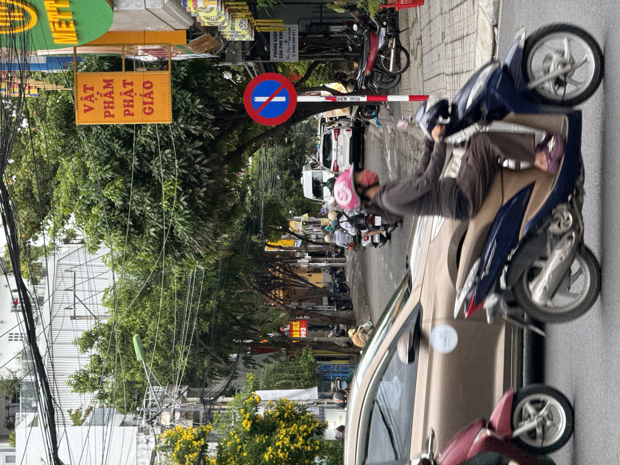 This image depicts a street scene in Nha Trang, Vietnam, characterized by a blend of urban elements and daily life. A gold-colored car is prominently positioned in the foreground, obscuring a portion of a red scooter. To the right, a person wearing a pink helmet and mask rides a dark blue scooter, their legs a blur of motion. Behind them, a white van is visible, with other vehicles and motorcycles further down the street.
The environment is a busy street lined with buildings, trees, and utility poles with a dense network of electrical wires overhead. Several signs are visible, including an orange sign with red Vietnamese text and a number, a blue and red prohibition sign indicating no parking, and a green sign with yellow lettering partially obscured by trees. There are also decorative yellow lanterns hanging from a building. The street is paved, and a sidewalk is visible on the right side, with a person sitting on the curb and a red basket near parked motorcycles. Yellow flowering trees add a splash of color to the left side of the frame. The overall impression is a snapshot of a typical day on a bustling Vietnamese city street.