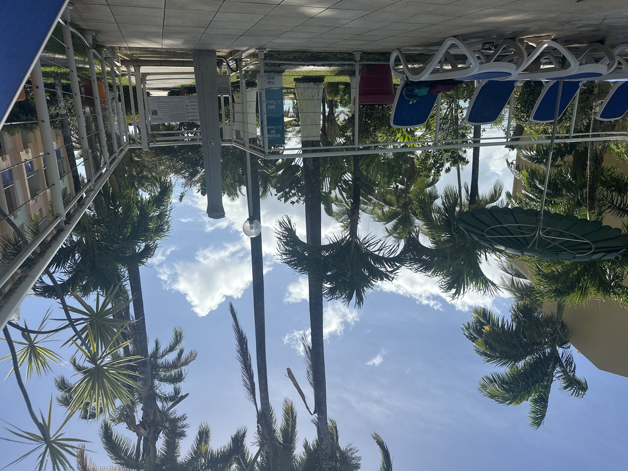 The image depicts a scene at a resort or hotel in Le Gosier, France. Several palm trees are visible against a blue sky with scattered clouds. In the foreground, there are lounge chairs with blue cushions arranged along a tiled patio or walkway. A metal railing and signage are also present, with text indicating “Plage” (Beach), “Snack”, and “Restaurant”. The scene appears to be near a pool or beach area, with tropical vegetation framing the view.