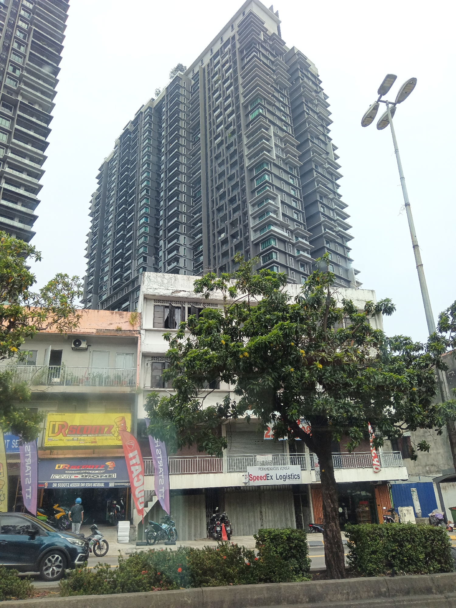 The image captures a street-level view in Kuala Lumpur, Malaysia, dominated by towering modern skyscrapers behind older, multi-story buildings. In the foreground, a dark blue sedan is parked on the left, with several motorcycles visible. A green tree stands prominently in the middle, partially obscuring the facades of the lower buildings. These buildings appear to house various businesses, including a motorcycle shop with prominent signage for "DR SCOOTZ AKSSERI DAN SERVIS MOTOSIKAL," and a logistics company, "SpeedEx Logistics." The lower buildings have a mix of open and shuttered storefronts, with balconies and air conditioning units visible on the upper floors. A tall street lamp with four lights stands to the right of the skyscrapers. The sky is overcast, suggesting a hazy or cloudy day.