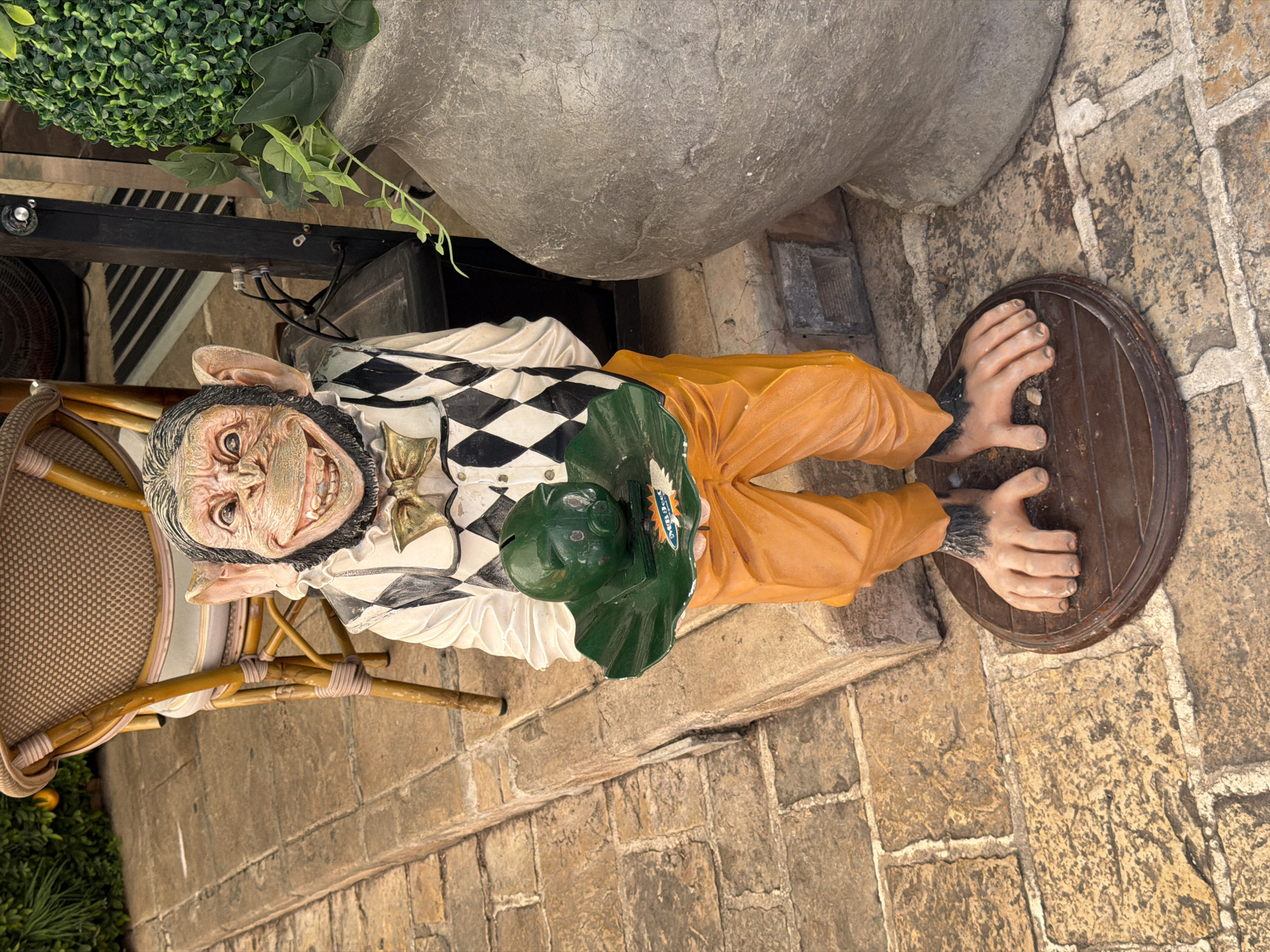 A life-sized, anthropomorphic chimpanzee statue stands on a circular wooden base on a paved stone patio. The statue is dressed in an outfit consisting of an orange jumpsuit, a white shirt with a ruffled collar, a black and white diamond-patterned vest, and a golden bow tie. It holds a green tray in its hands, upon which sits a green piggy bank shaped like a pig's head. The statue has a realistic, albeit exaggerated, chimpanzee face with a wide, toothy grin and large ears. The background includes a wicker chair, a large grey planter with greenery, and a hint of a building facade. The overall scene appears to be an outdoor decorative display, possibly in a restaurant or garden setting. The location is specified as Budva, Montenegro.