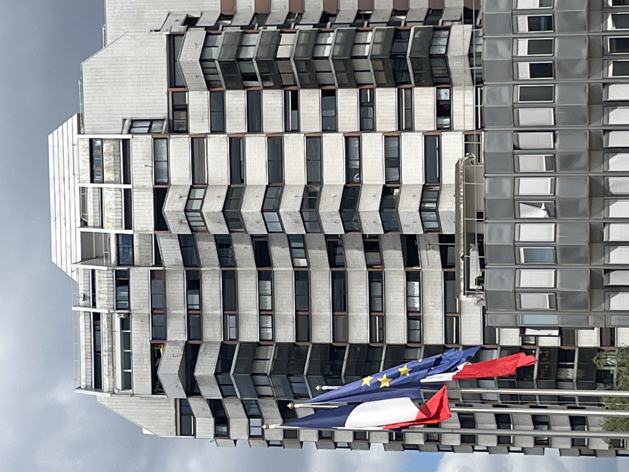 The image shows a modern, multi-story building with a distinctive stepped facade and numerous windows, some with balconies. In the foreground, three flags are visible: the French flag, the European Union flag, and another flag, possibly red. The flags are on flagpoles, with the French flag and the EU flag prominently displayed. The building appears to be in an urban setting, with a cloudy sky overhead. This building is located in Créteil, France.