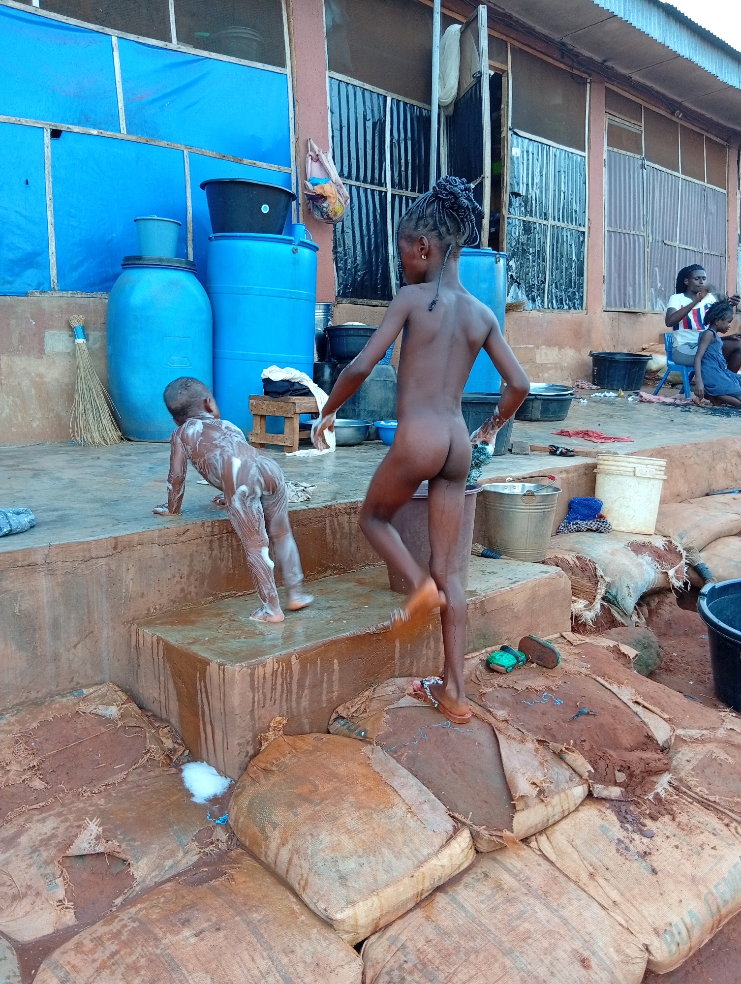 In Ogidi, Nigeria, two children are outside a building. One child, covered in soap, is crawling up cement steps. The other, wearing sandals, is walking down the steps. Blue barrels are stacked behind them. Adults and other children are in the background. Bags are stacked on the ground. Buckets, basins, brooms, and sandals are scattered around. The building has a blue tarp and corrugated metal.
